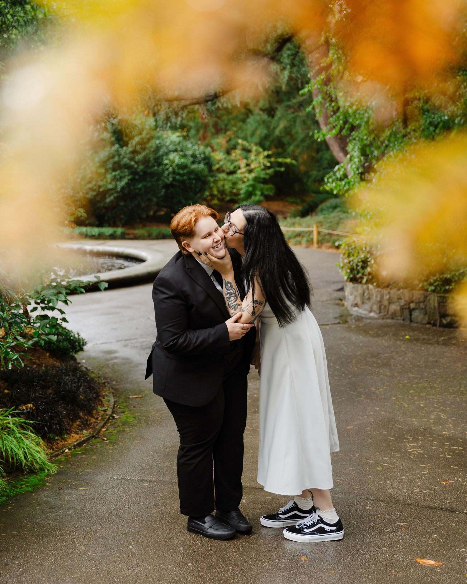Couple sharing a tender kiss in a lush garden path, framed by golden autumn foliage, one in black suit other in white dress.