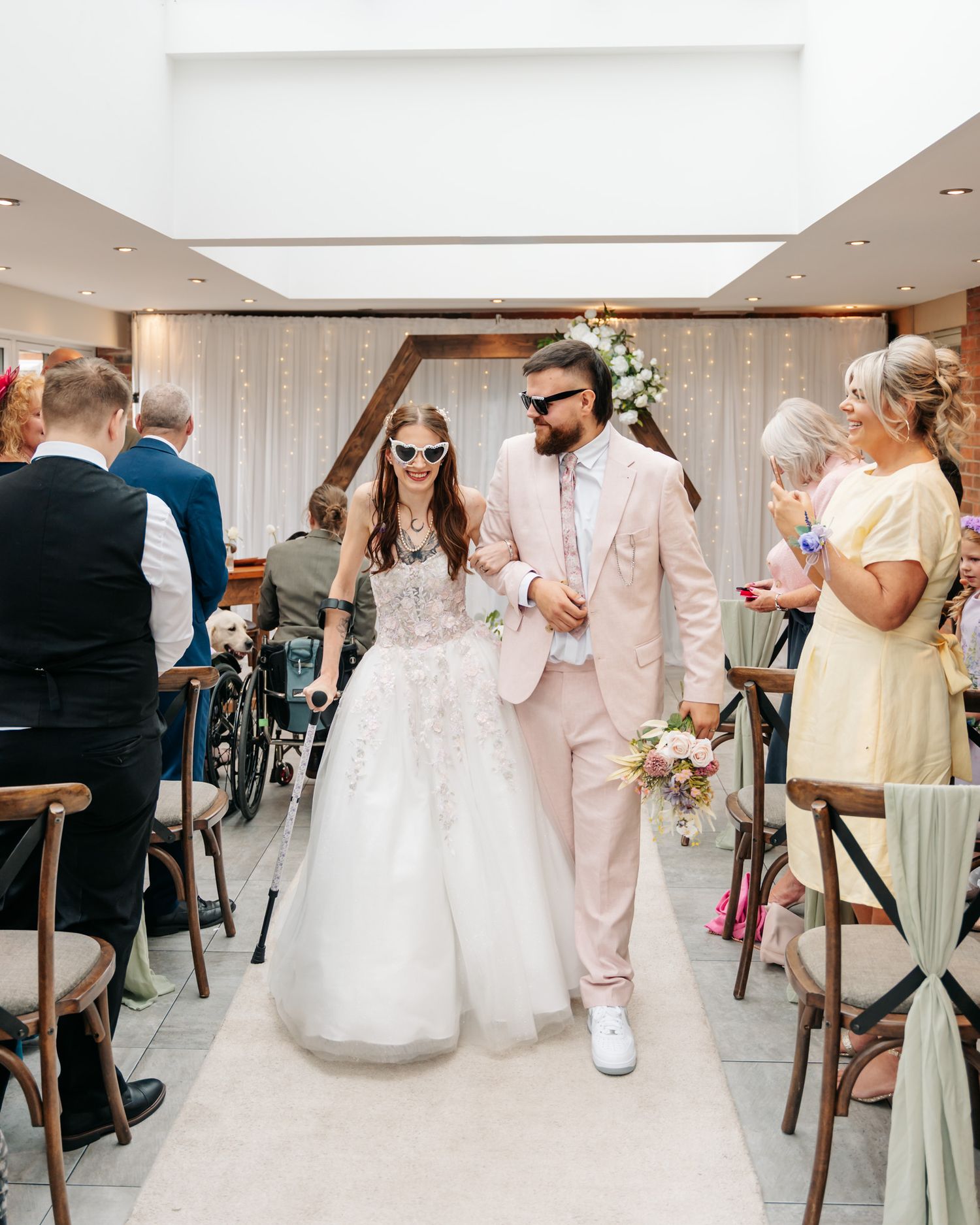 Newlyweds walk down the aisle wearing novelty sunglasses, bride in white gown, groom in pink suit, guests applauding.