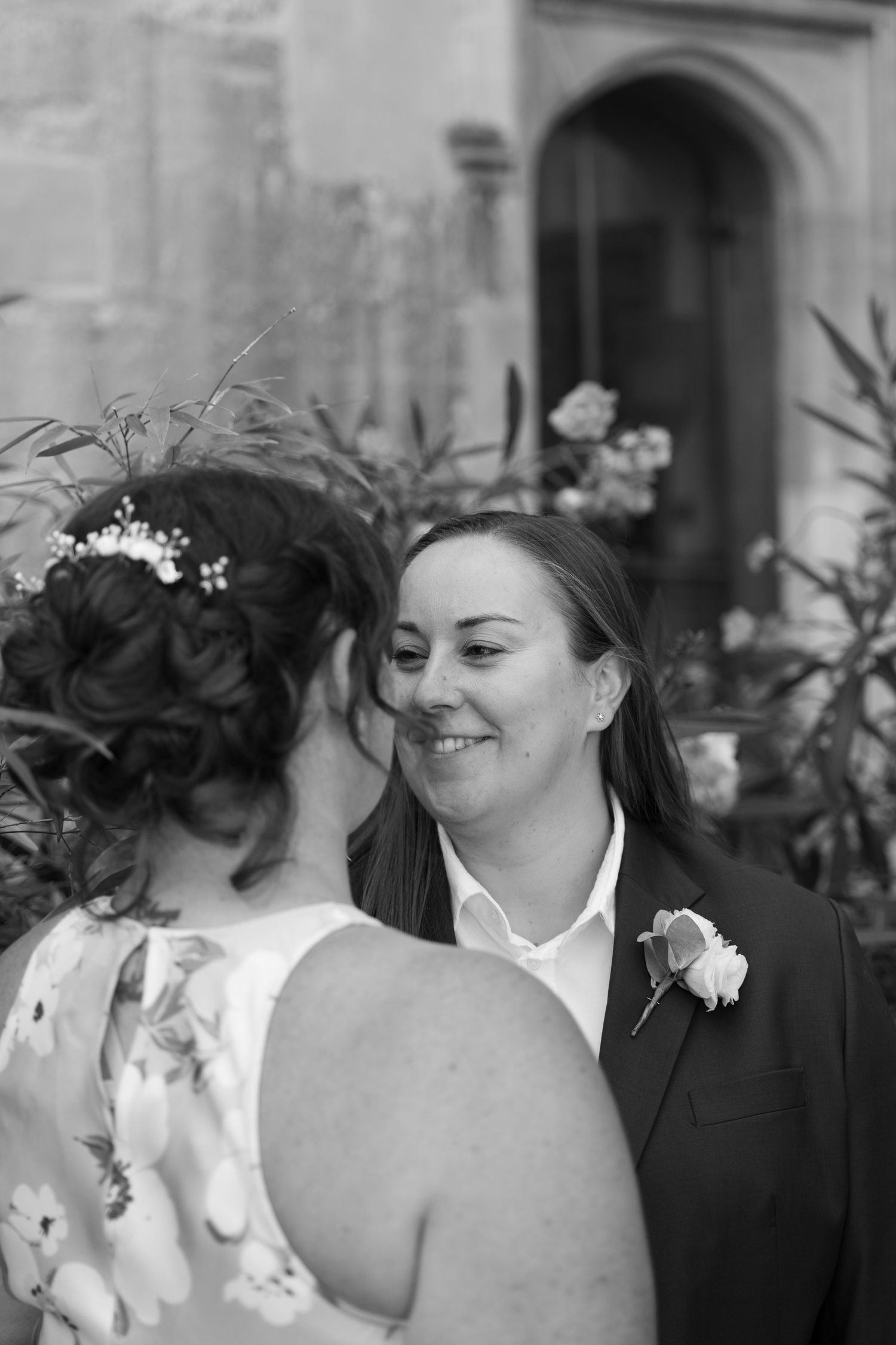 Black and white wedding photo of two brides smiling at each other, one wearing a suit with boutonniere.