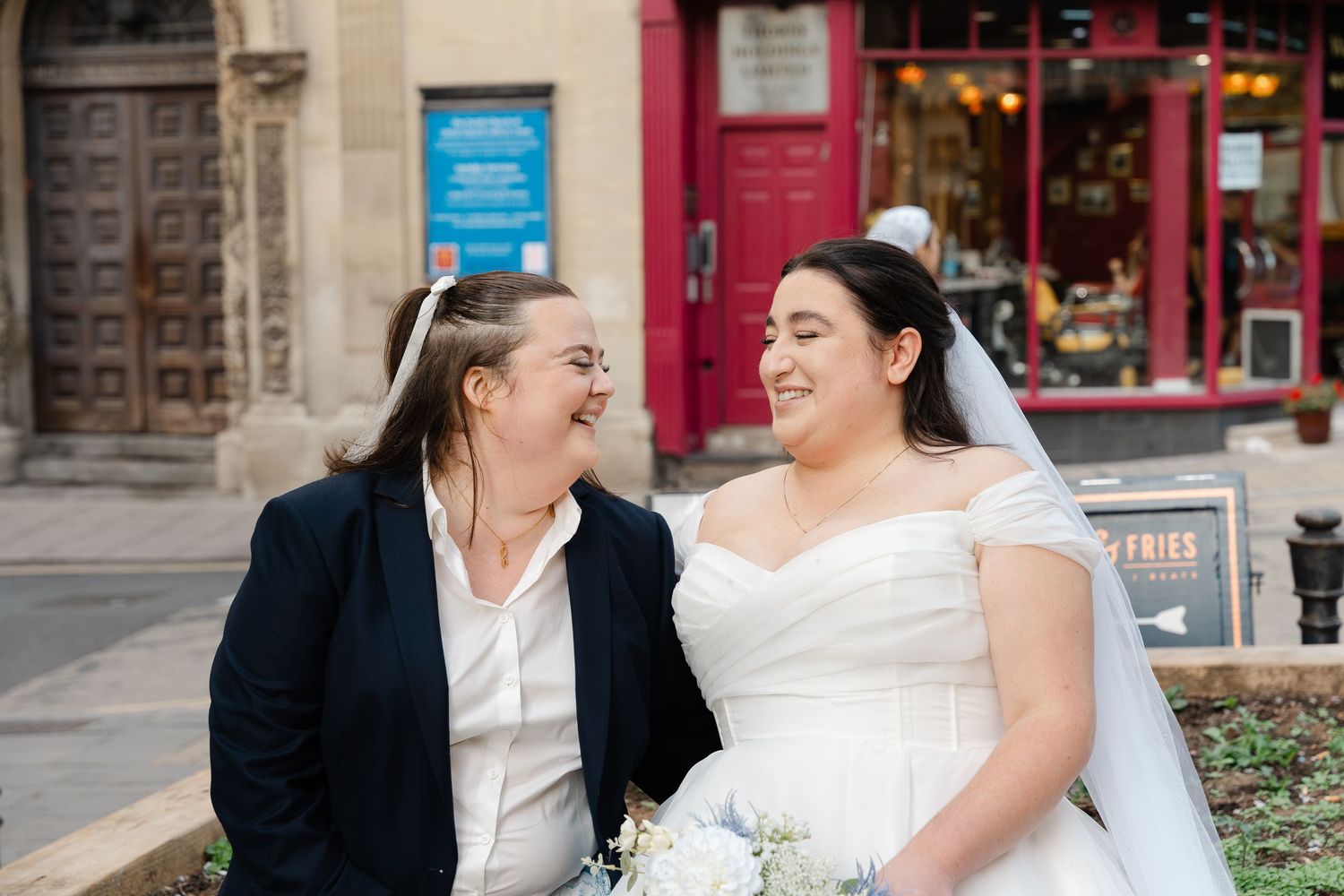 Two brides share a joyful laugh outdoors, one in a white off-shoulder gown with veil, the other in a navy blazer.
