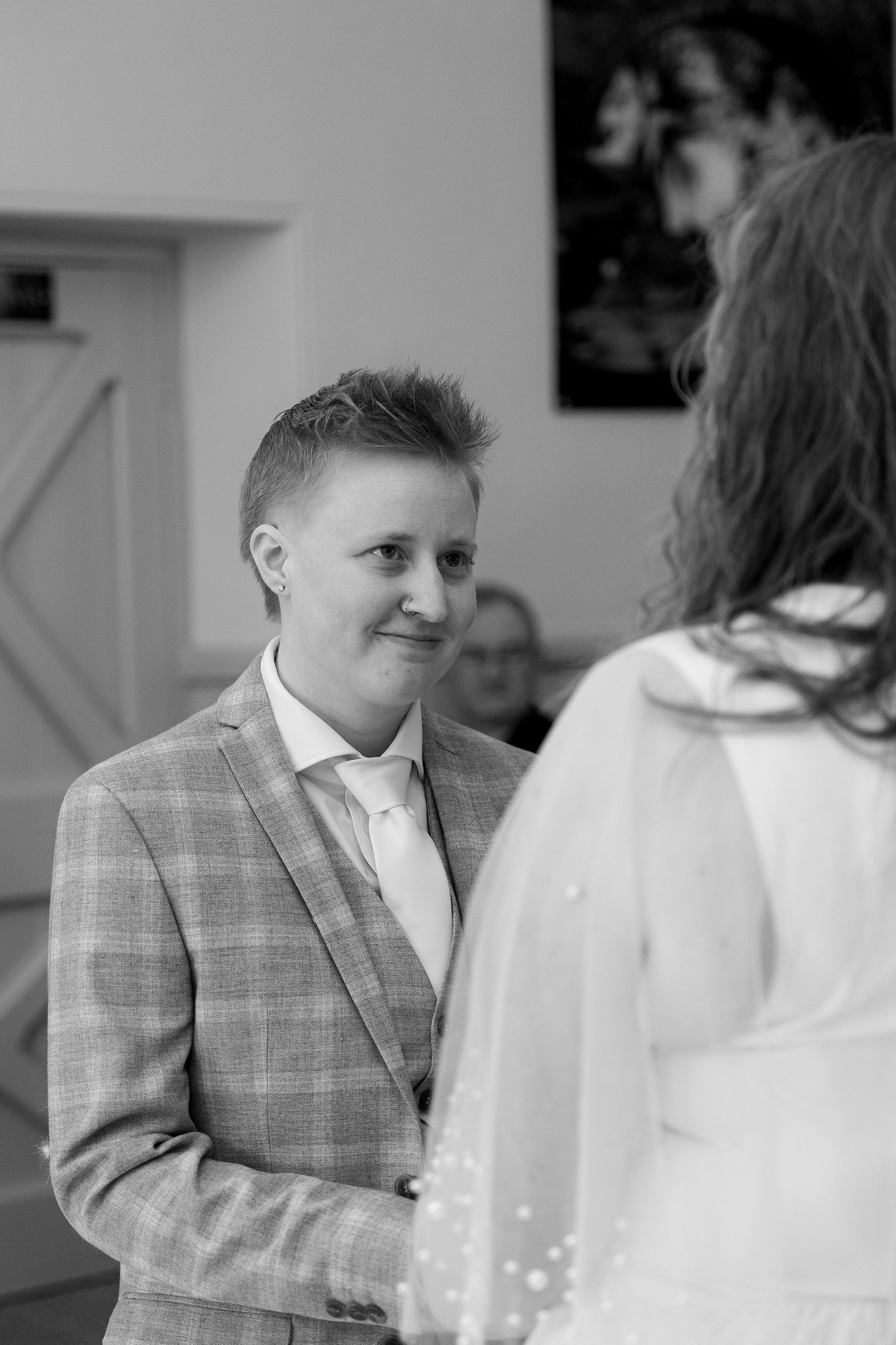 Black and white wedding photo of a person in a plaid suit smiling warmly at their partner in a white dress.