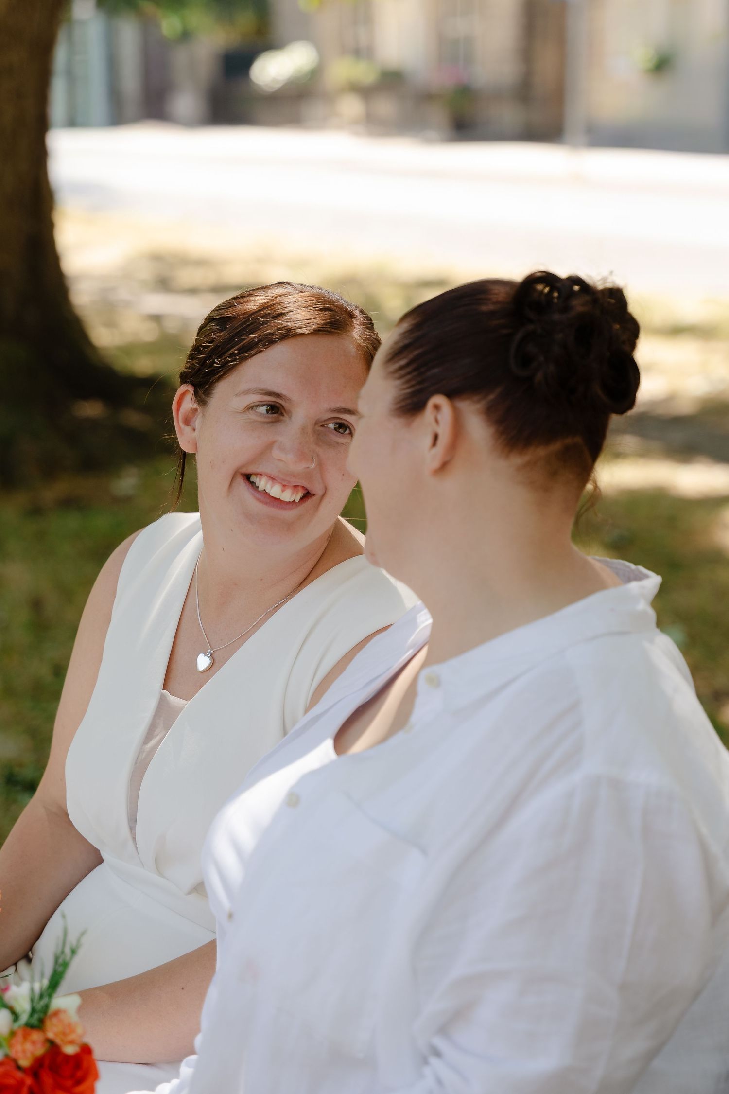 Two women in white attire share a joyful moment outdoors, one smiling warmly while holding orange flowers.