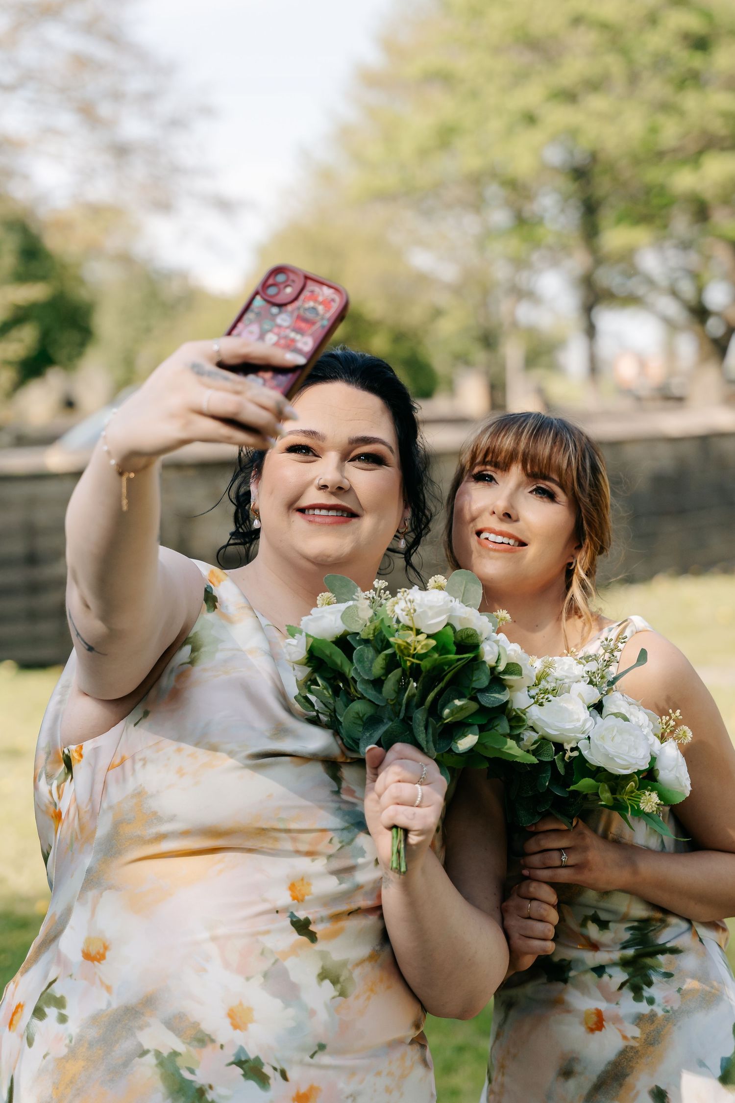 Two bridesmaids in floral dresses take a selfie outdoors, one holding a white bouquet of flowers at a wedding.