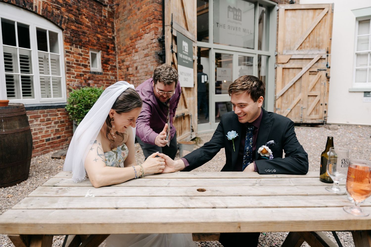 Bride and groom laughing with guest at outdoor wooden picnic table during wedding reception at rustic brick venue.