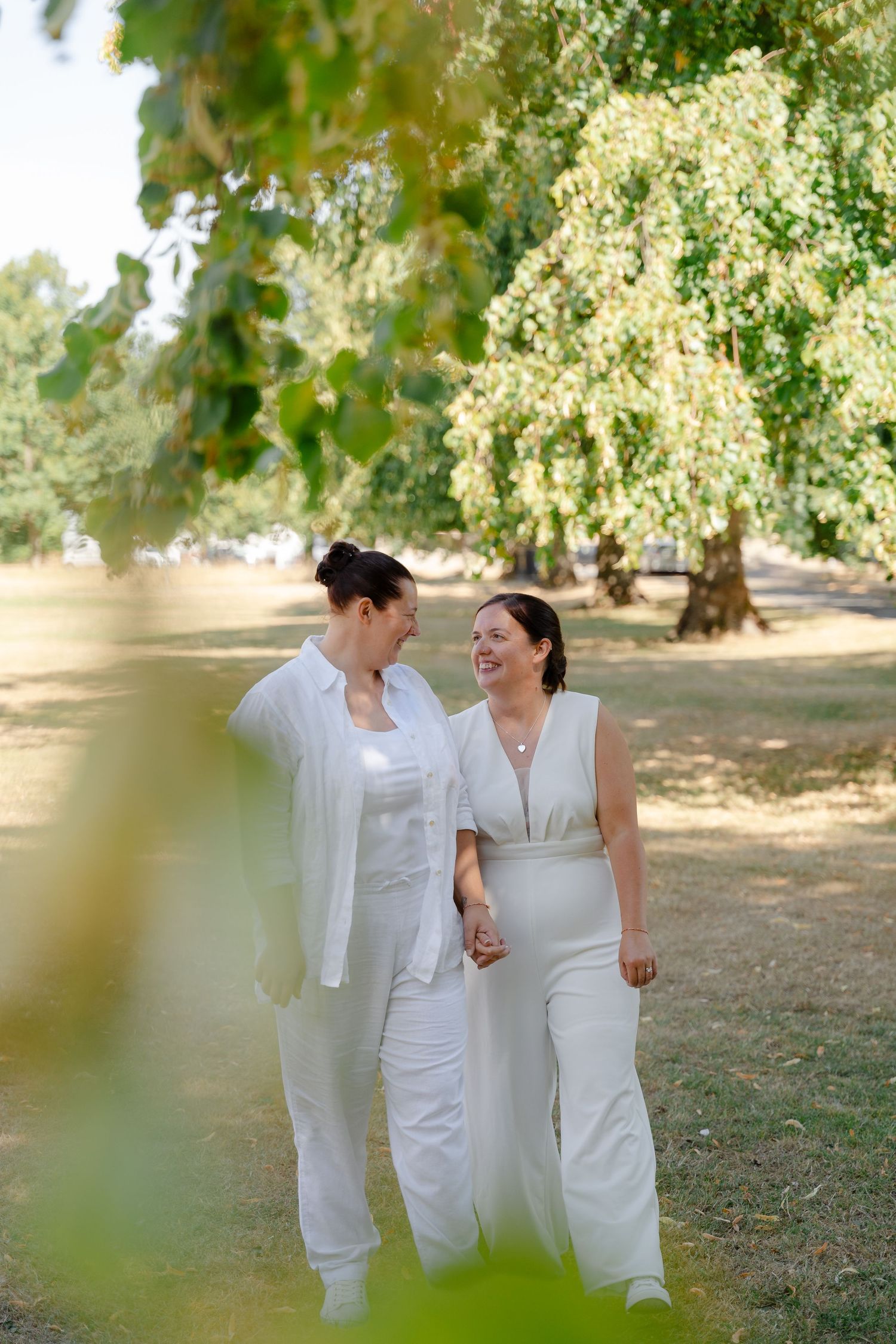 Two women in white outfits hold hands and smile while walking through a sunny park with lush green trees.