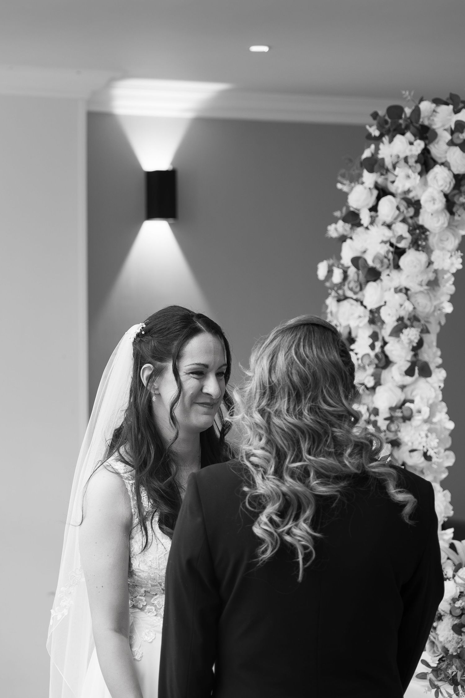 Black and white photo of a smiling bride in veil exchanging vows with partner before a floral arch at wedding ceremony.