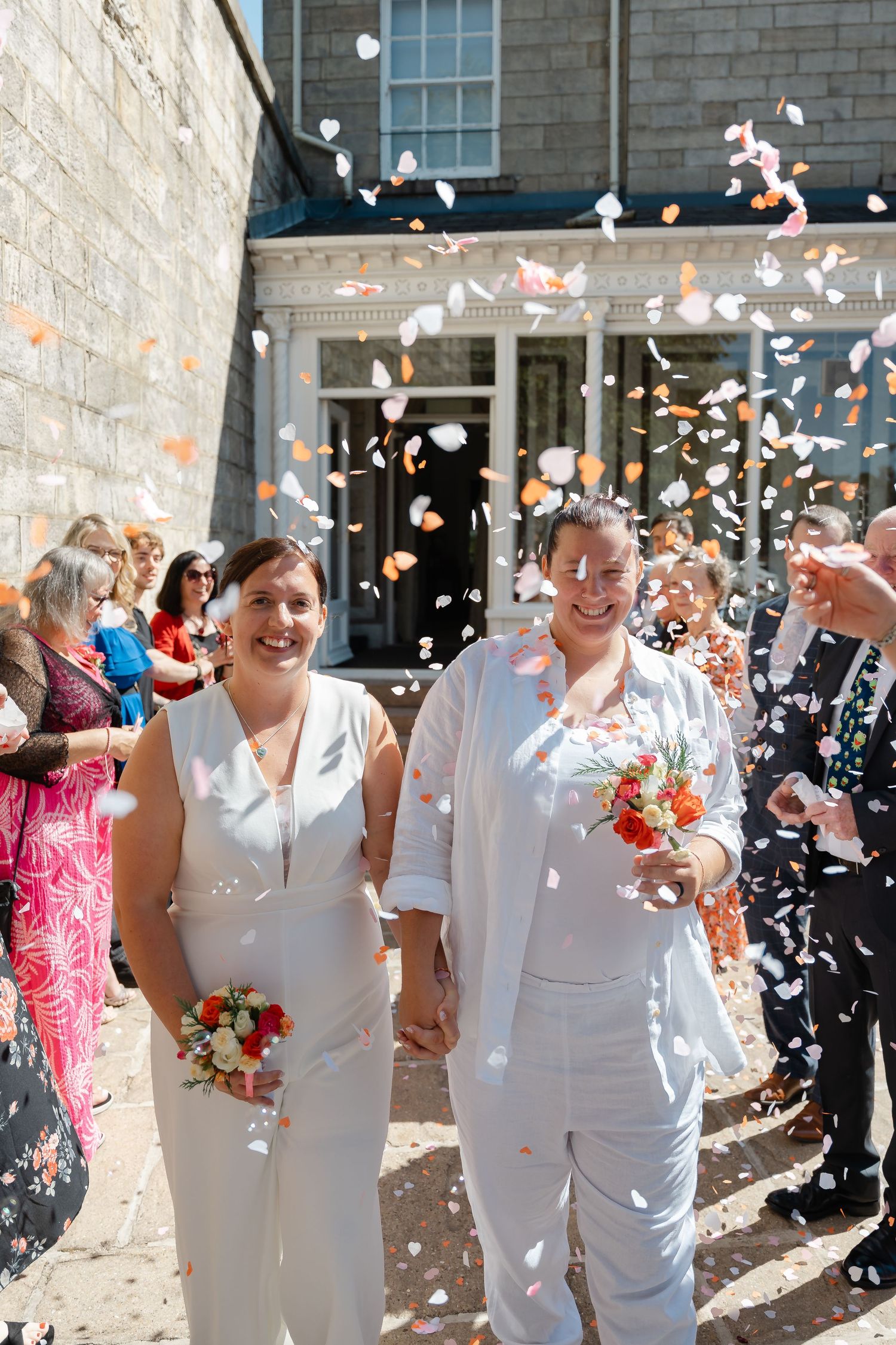 Two brides in white suits walk hand-in-hand through a shower of orange and white confetti outside a stone building.