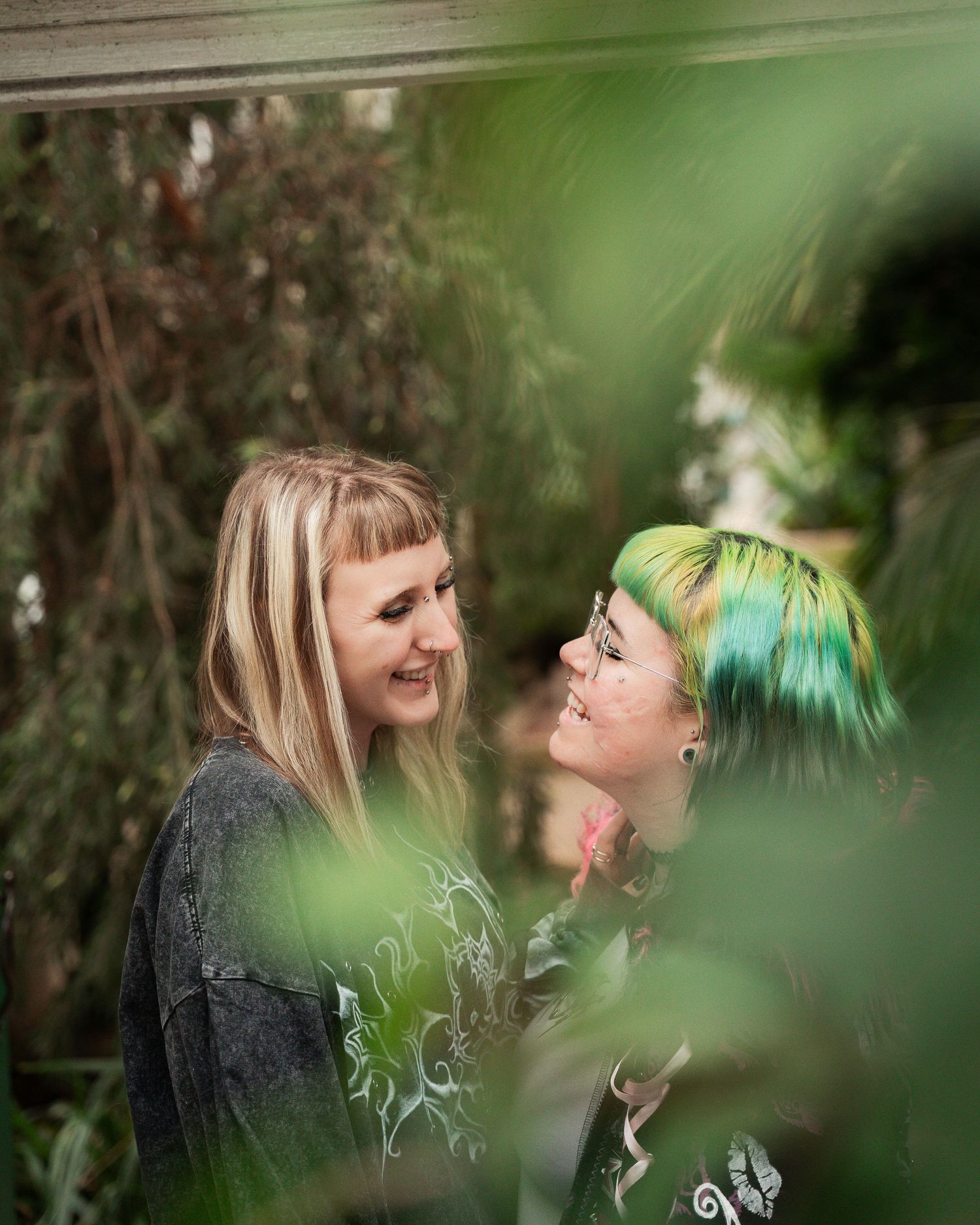 Two women smiling at each other outdoors, one with blonde hair and one with vibrant green hair, framed by lush green foliage.