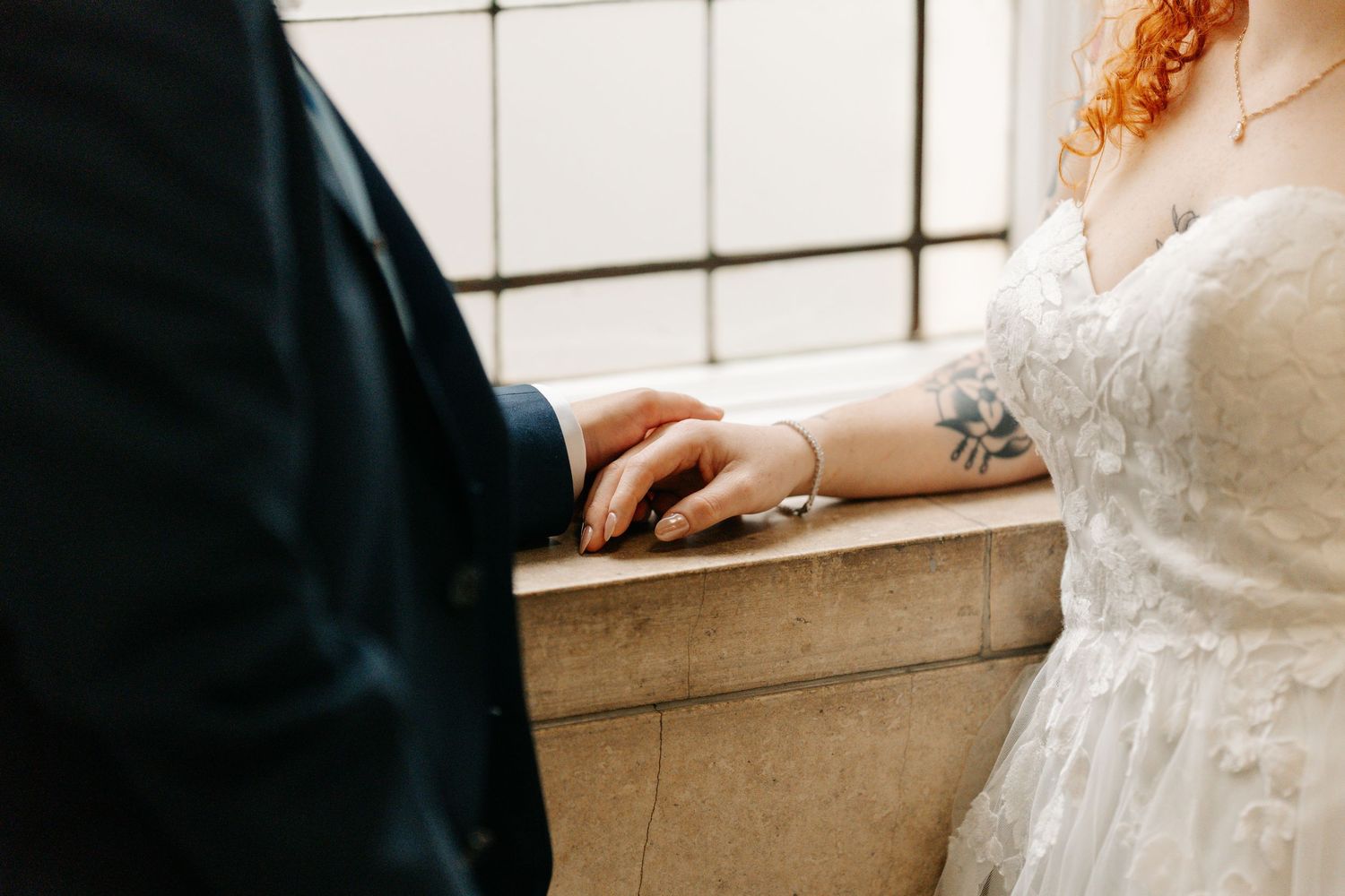 Bride and groom touching hands on stone ledge, bride wearing lace dress with tattoo and bracelet visible.