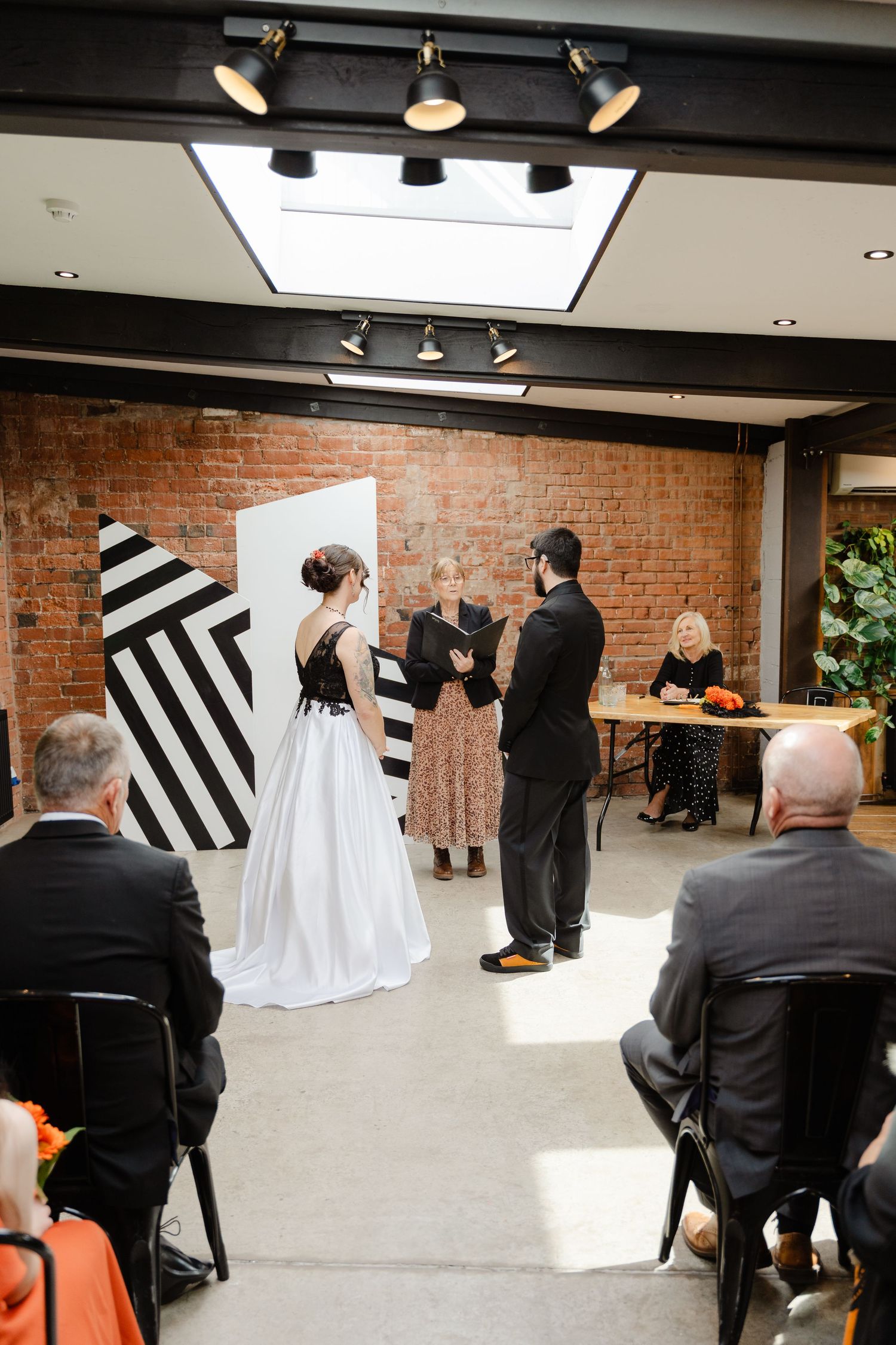 A bride and groom exchange vows at an indoor wedding ceremony with brick walls and a bold black-and-white geometric backdrop.