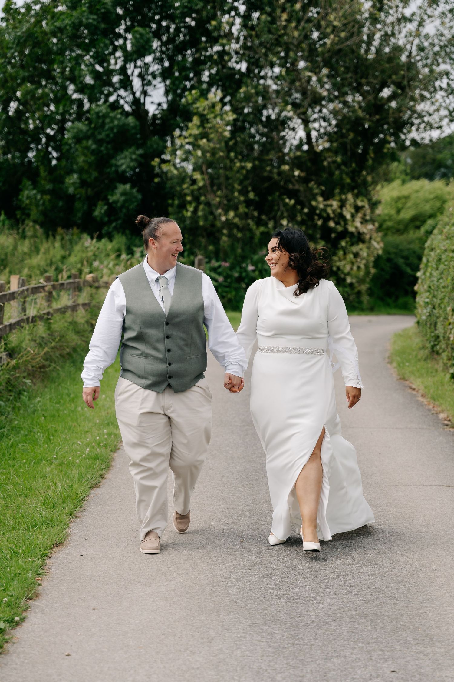 A couple in wedding attire holds hands and walks along a tree-lined path, smiling at each other on their wedding day.