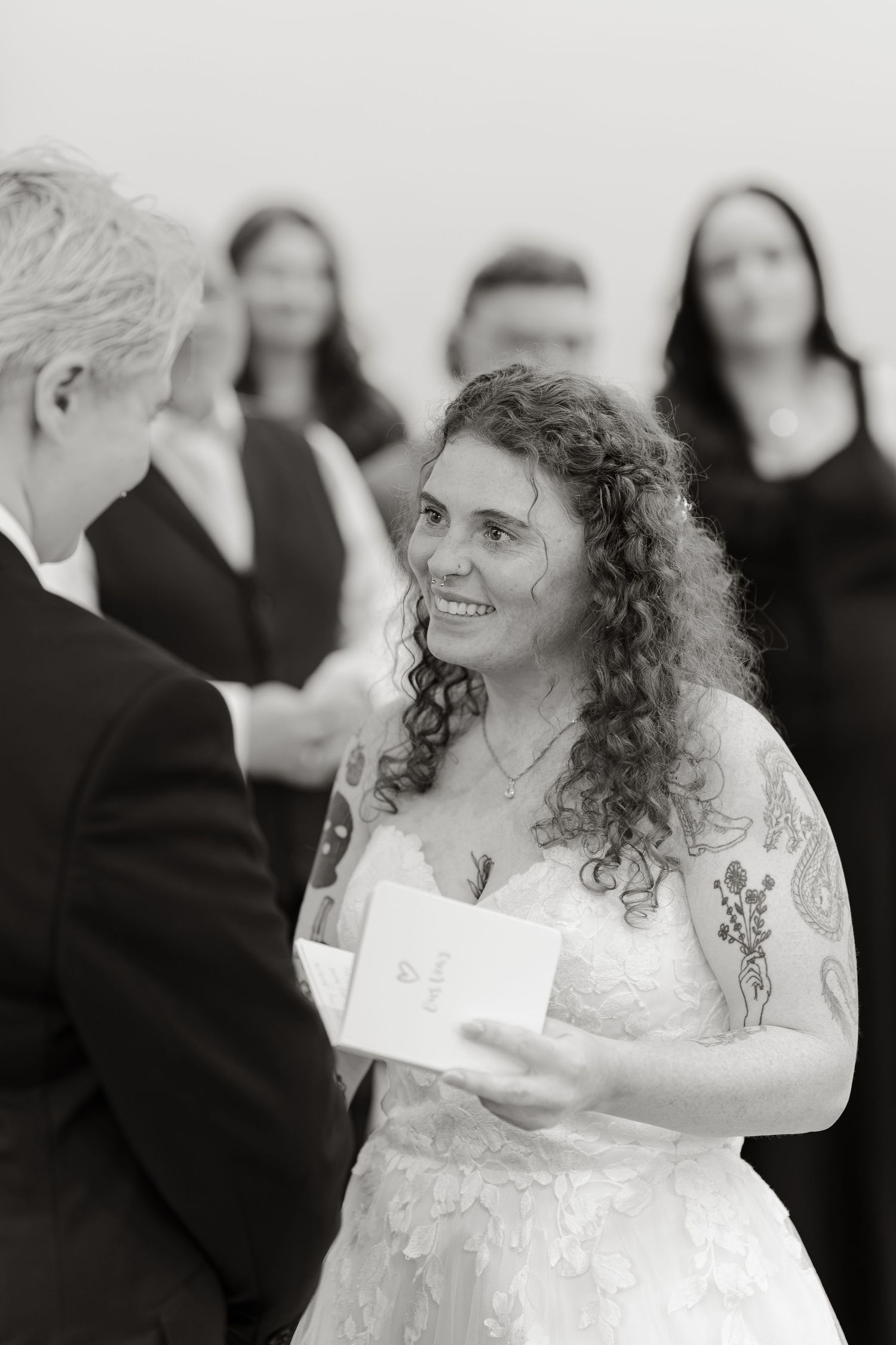 Bride with curly hair smiling during wedding ceremony, holding vows card, in elegant lace dress, black and white photo.