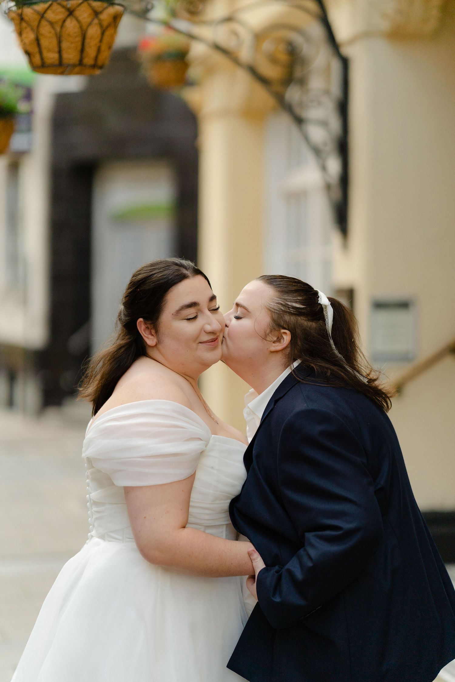 Two brides share a kiss, one in a white off-shoulder gown and one in a navy suit, on their wedding day.