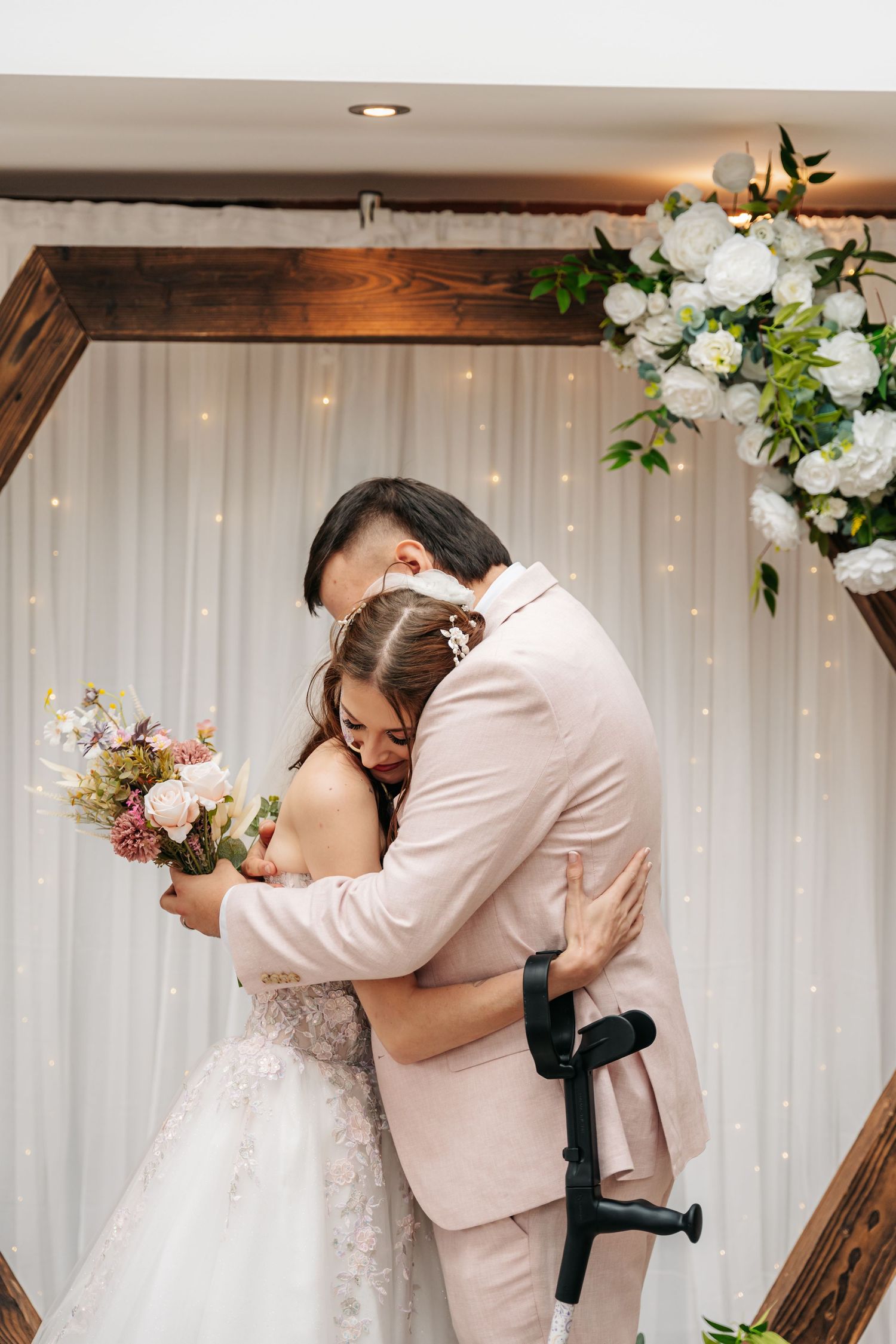 Bride and groom share an emotional first hug at altar decorated with white floral arch and fairy lights.