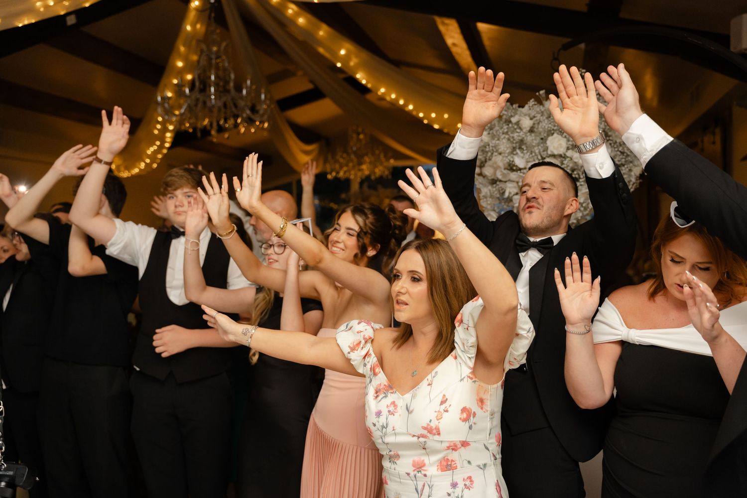 Guests dancing with hands raised at an elegant wedding reception with warm string lights.