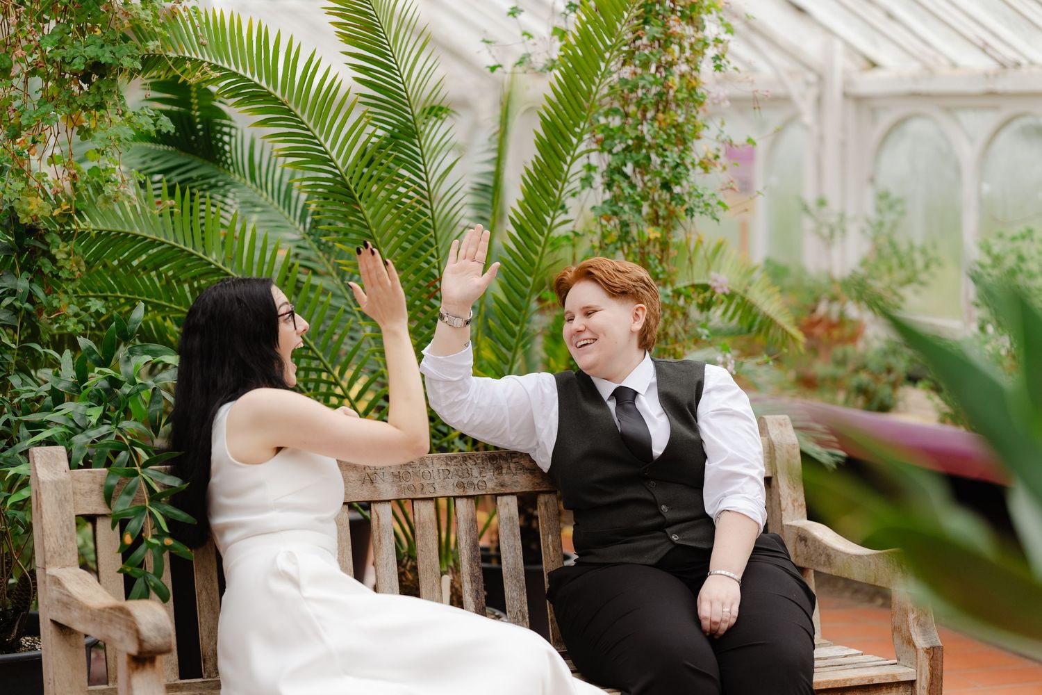 Two brides high-fiving joyfully in a lush greenhouse, one in a white gown and one in a black vest and tie.