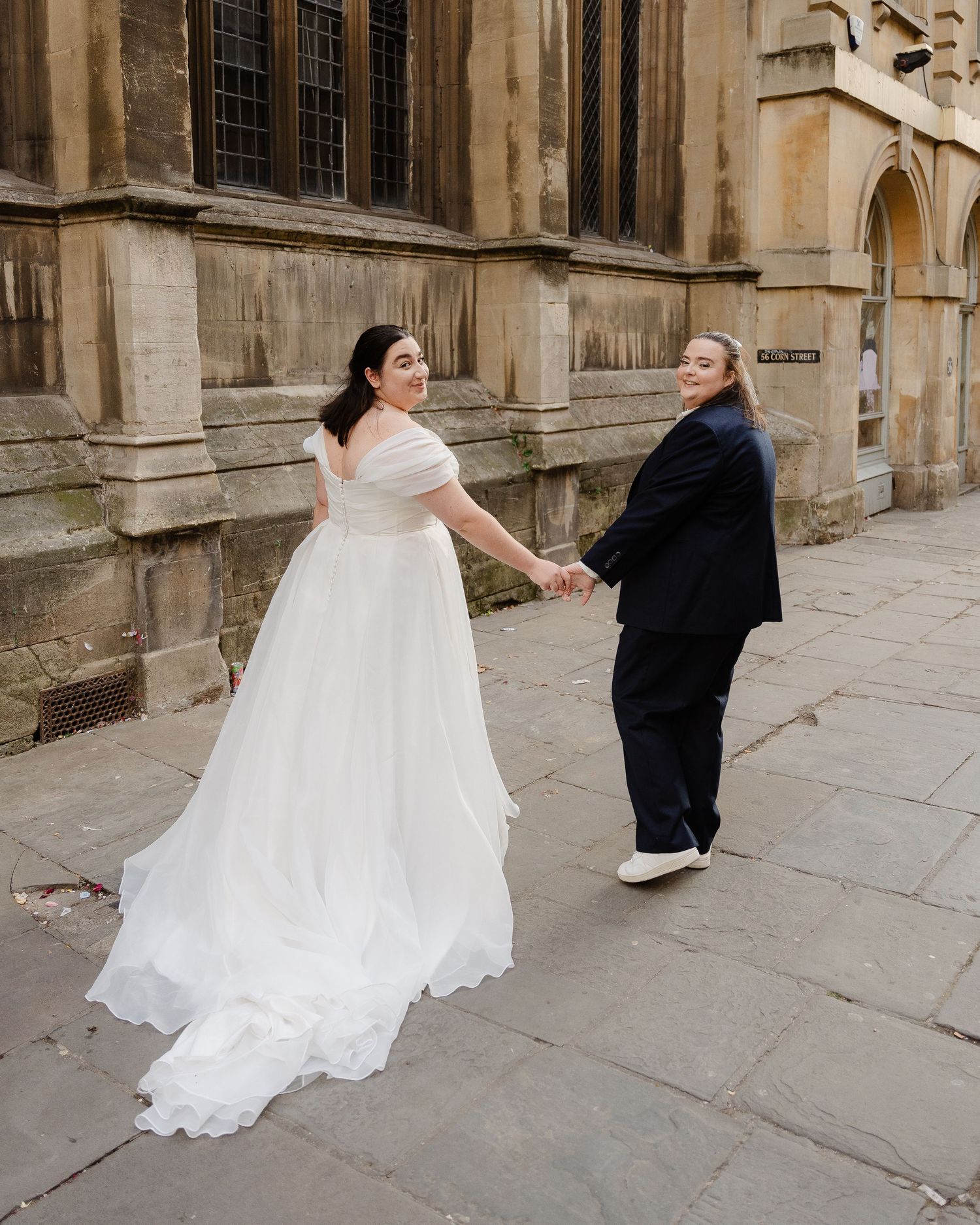 Two brides hold hands outside a historic stone building, one in a white off-shoulder gown, the other in a navy suit.