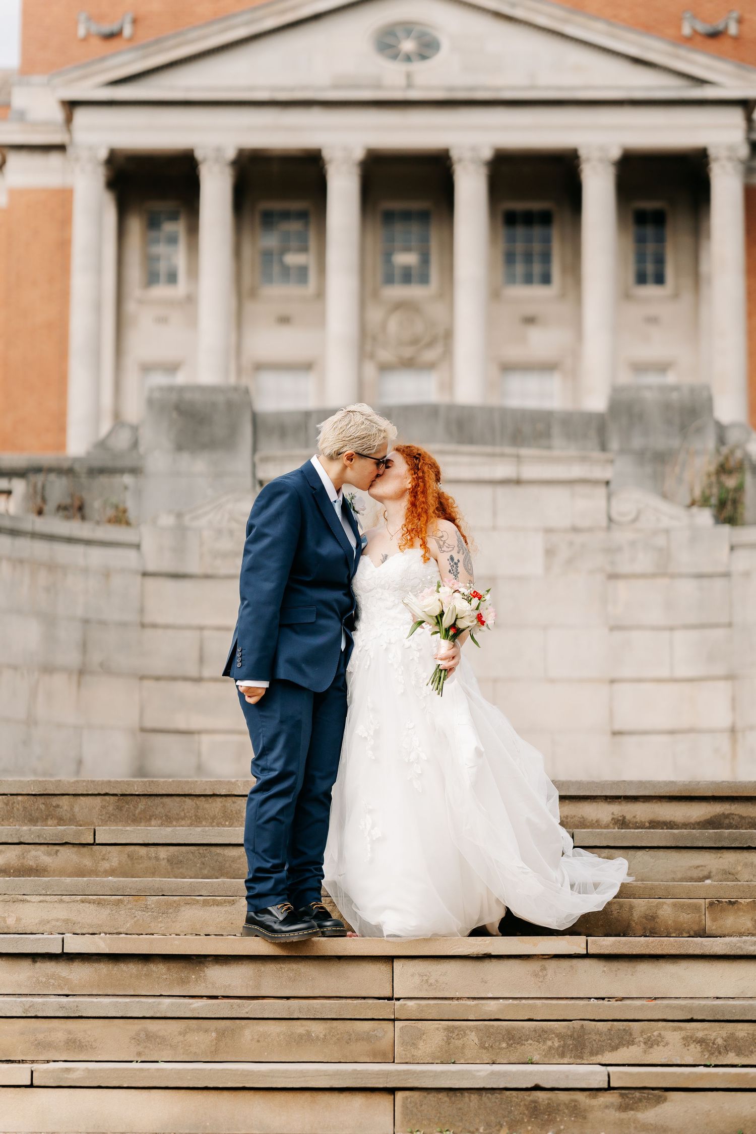 Bride and groom share a kiss on stone steps outside a classical building with columns on their wedding day.