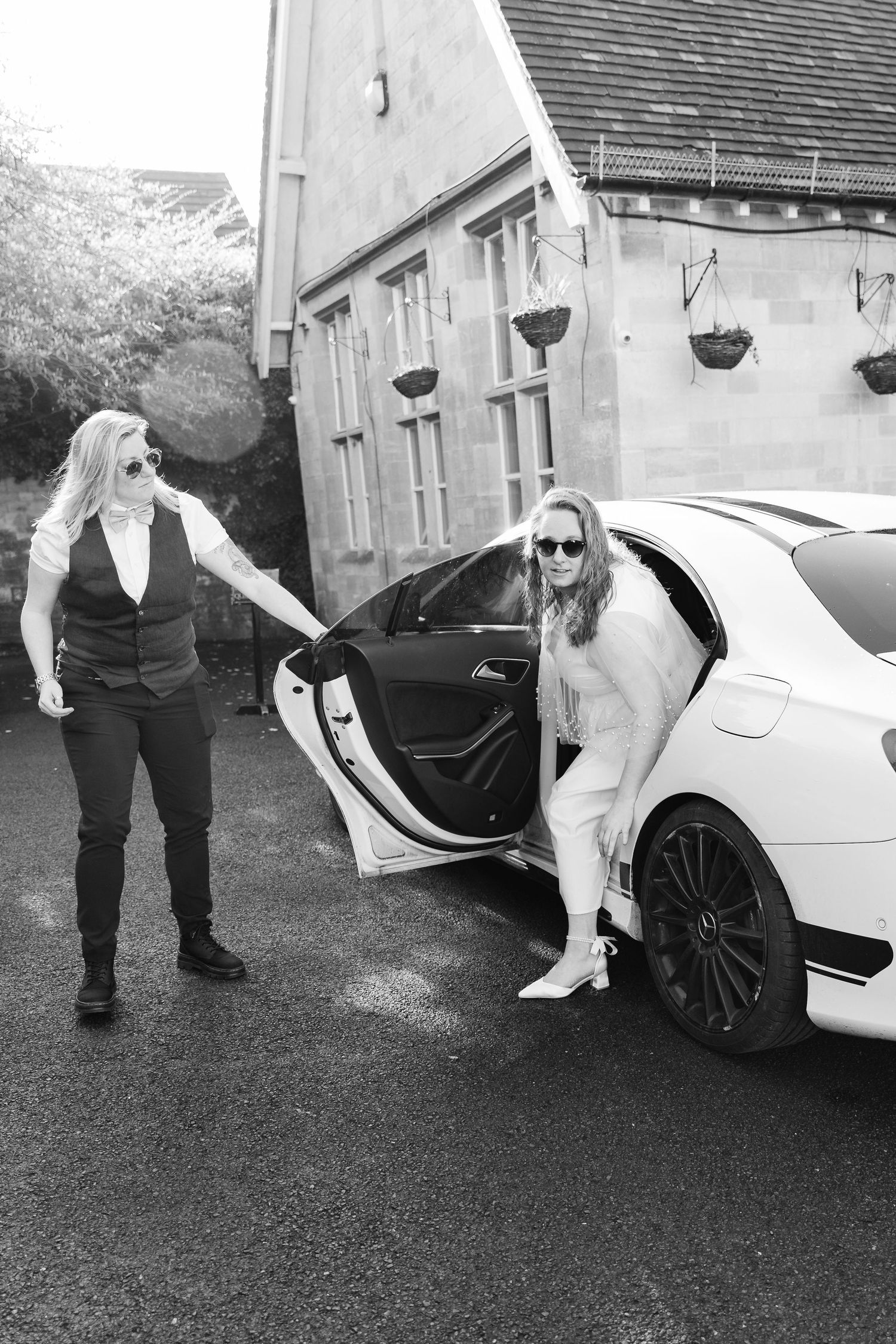 Black and white photo of two women in sunglasses, one holding open a white luxury car door outside a stone building.
