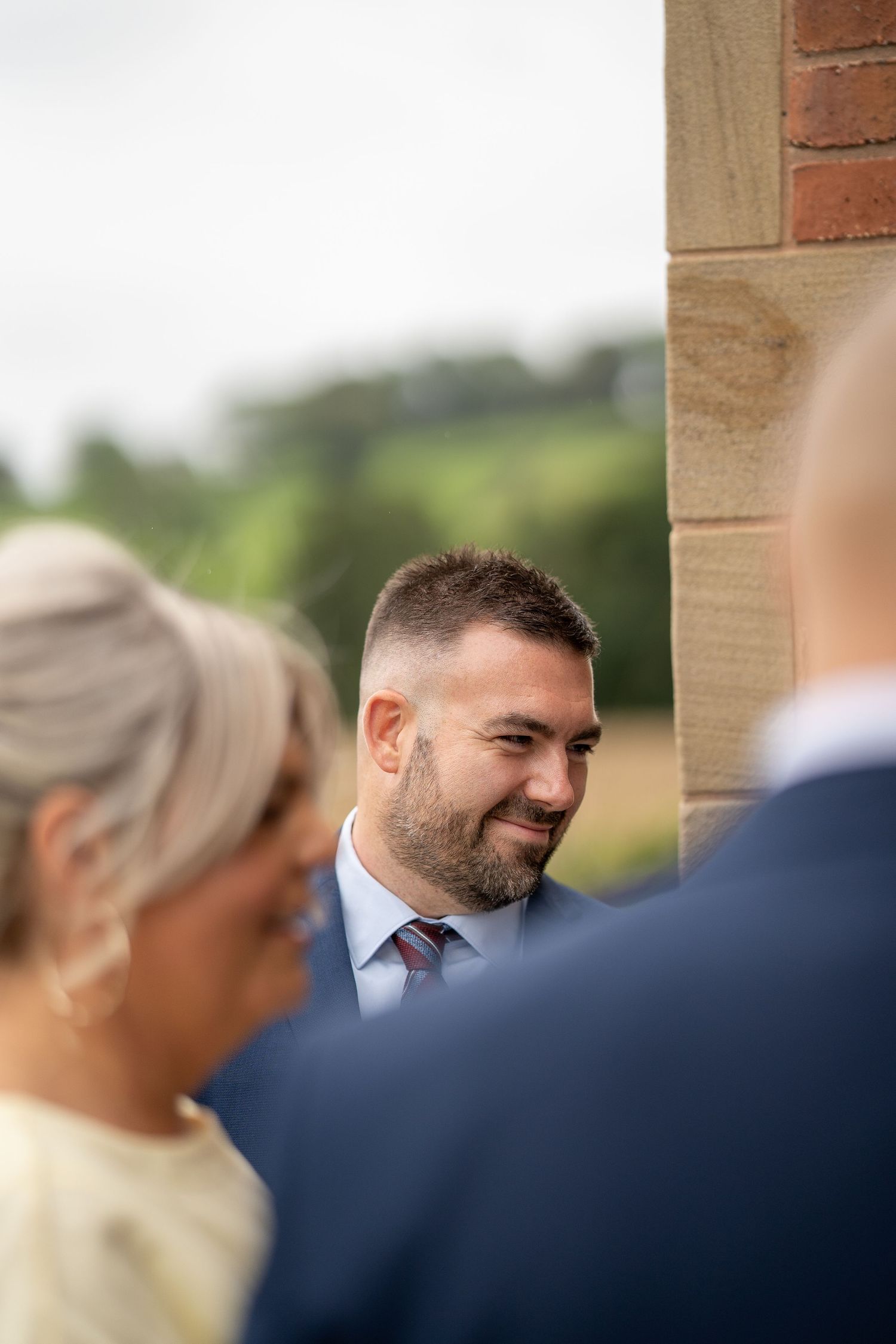 A bearded groom smiles warmly at a wedding ceremony outdoors, with guests in the foreground and greenery behind.