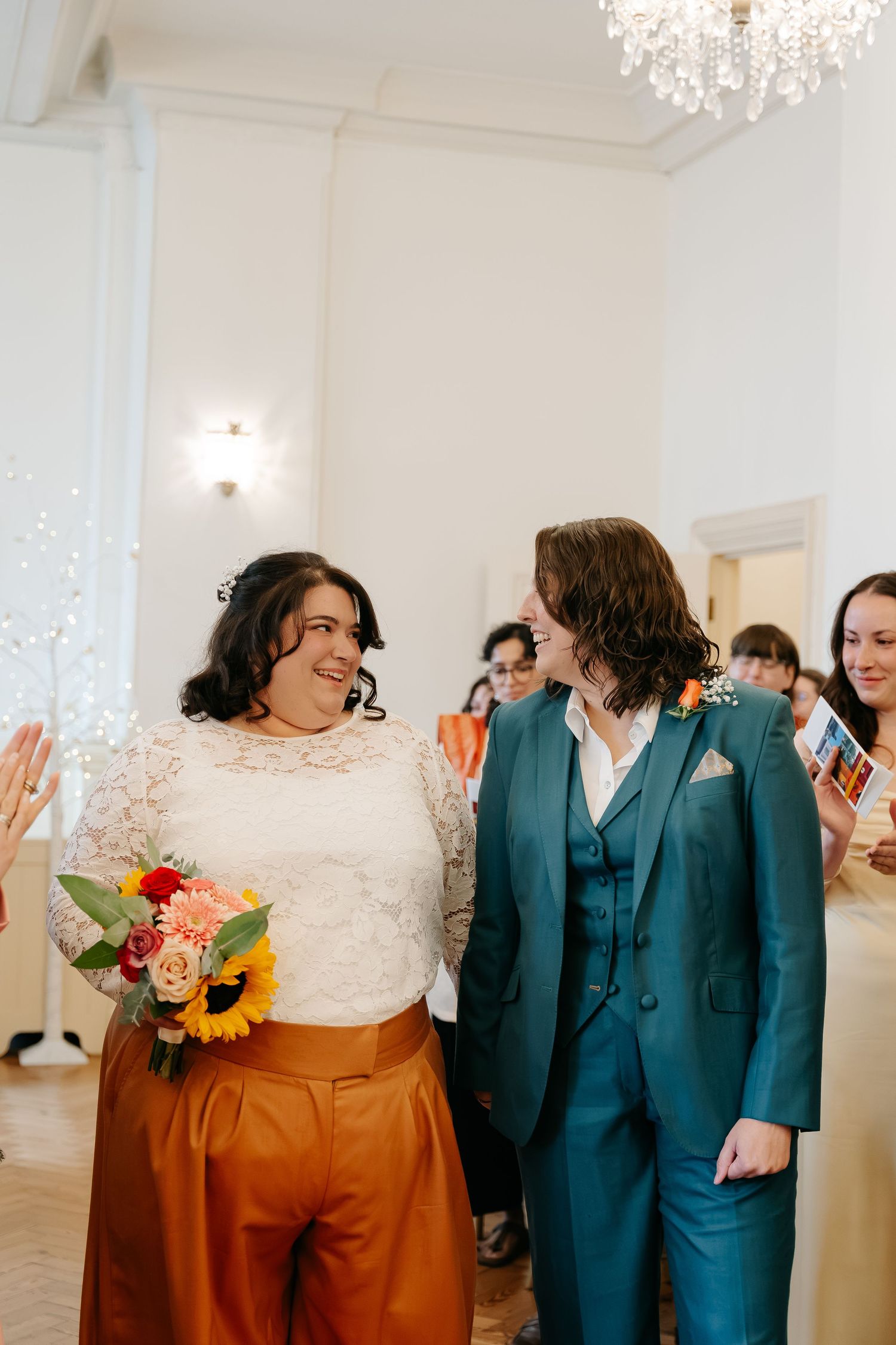 Two brides walk down the aisle, one in lace top with orange pants holding sunflower bouquet, other in teal suit, smiling joyfully.