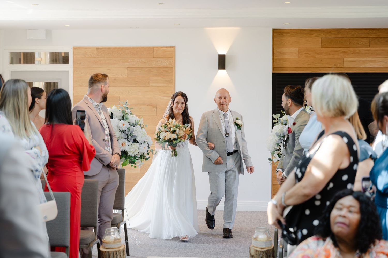 Bride walks down the aisle with her father during an elegant indoor wedding ceremony, guests seated on either side.