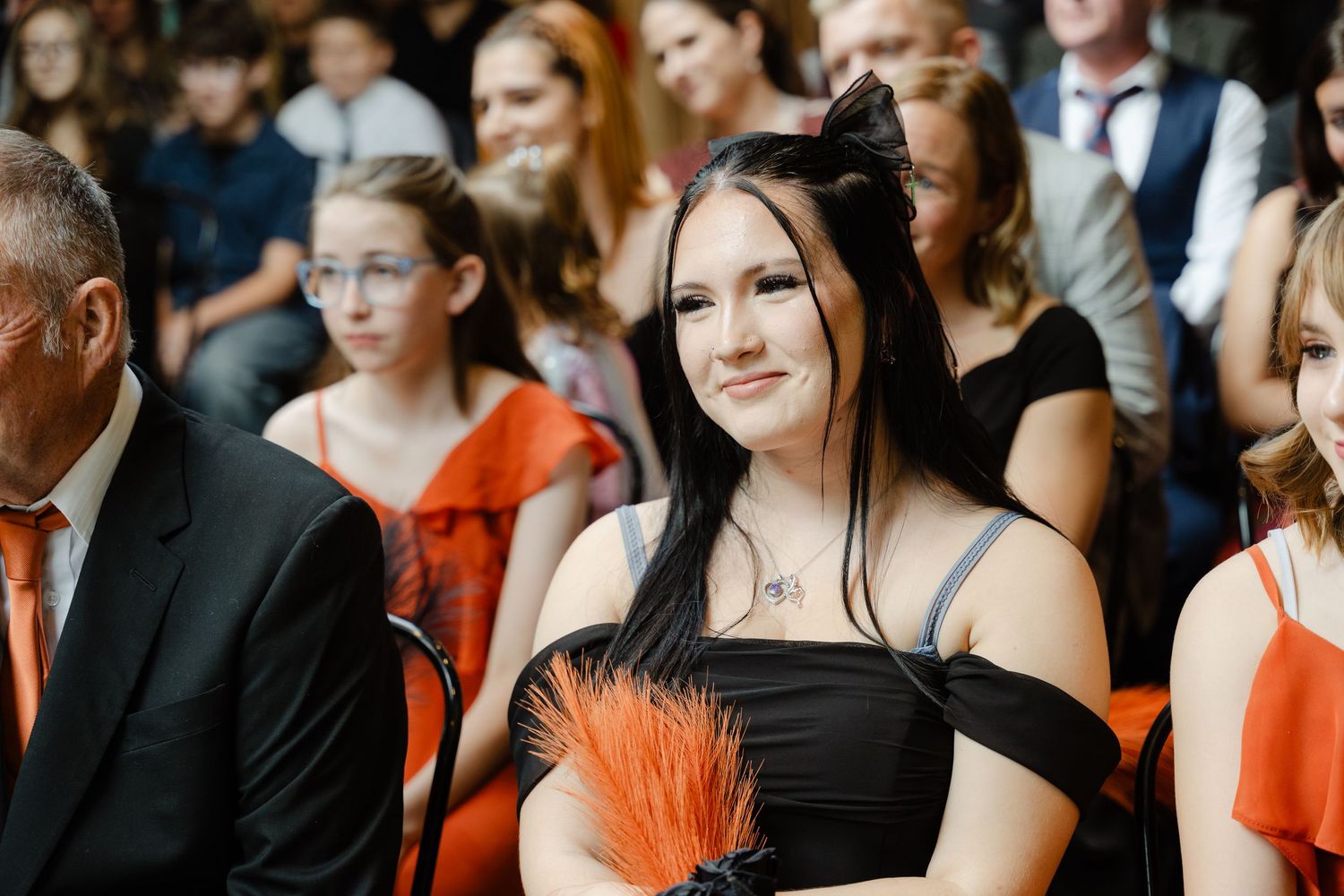 Young woman in black off-shoulder dress with orange feather accessory smiling at a formal event.
