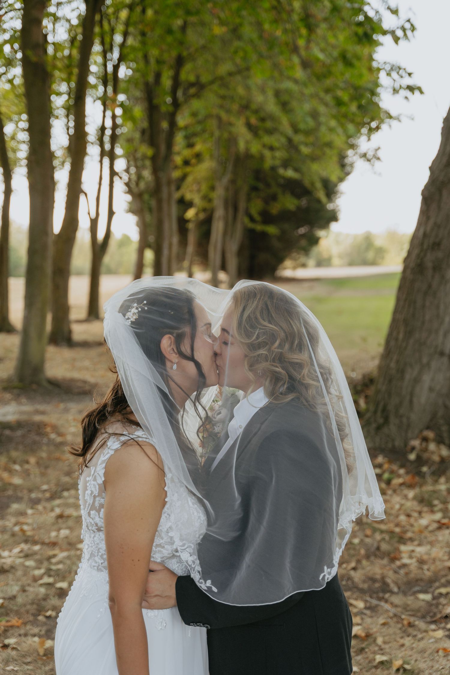 Two brides sharing a kiss under a wedding veil outdoors among tall trees on their wedding day.