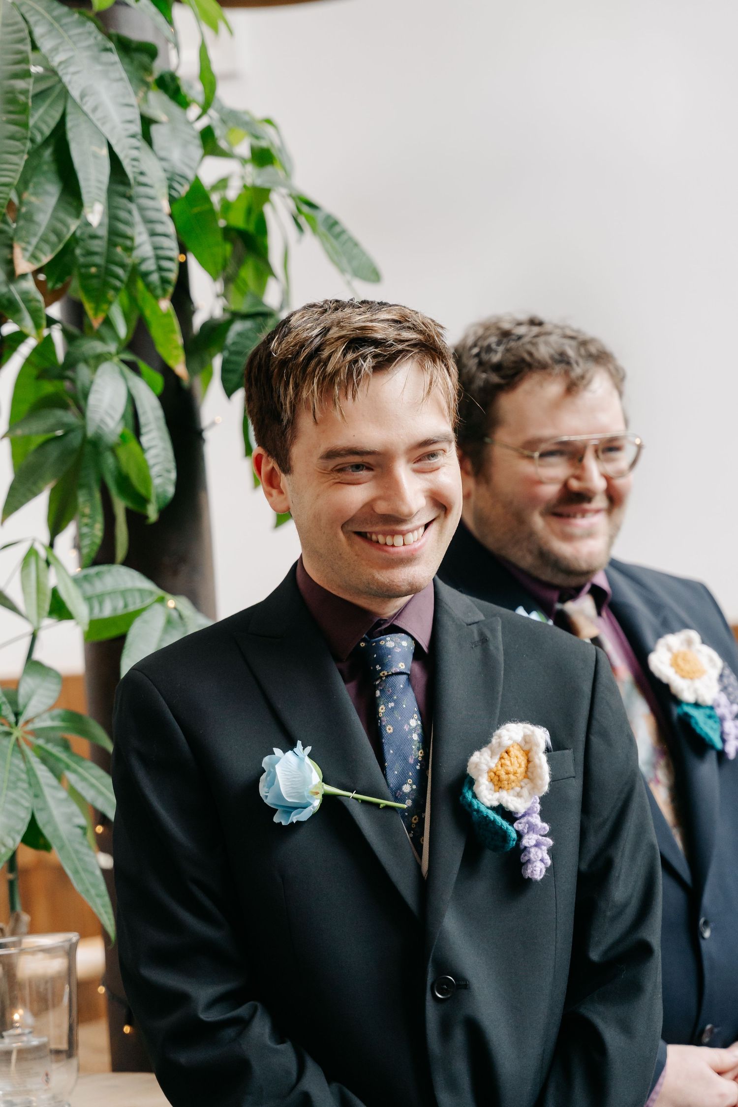 Two groomsmen in dark suits with floral boutonnieres smiling during a wedding ceremony indoors.