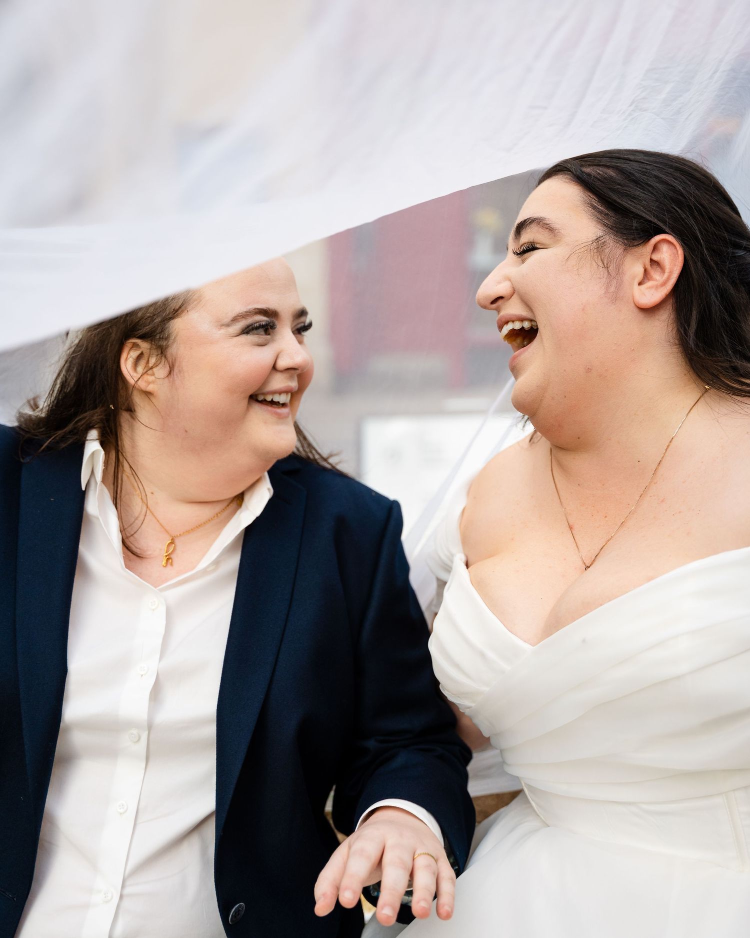 Two brides laughing joyfully under a wedding veil, one in a navy suit and one in a white dress.