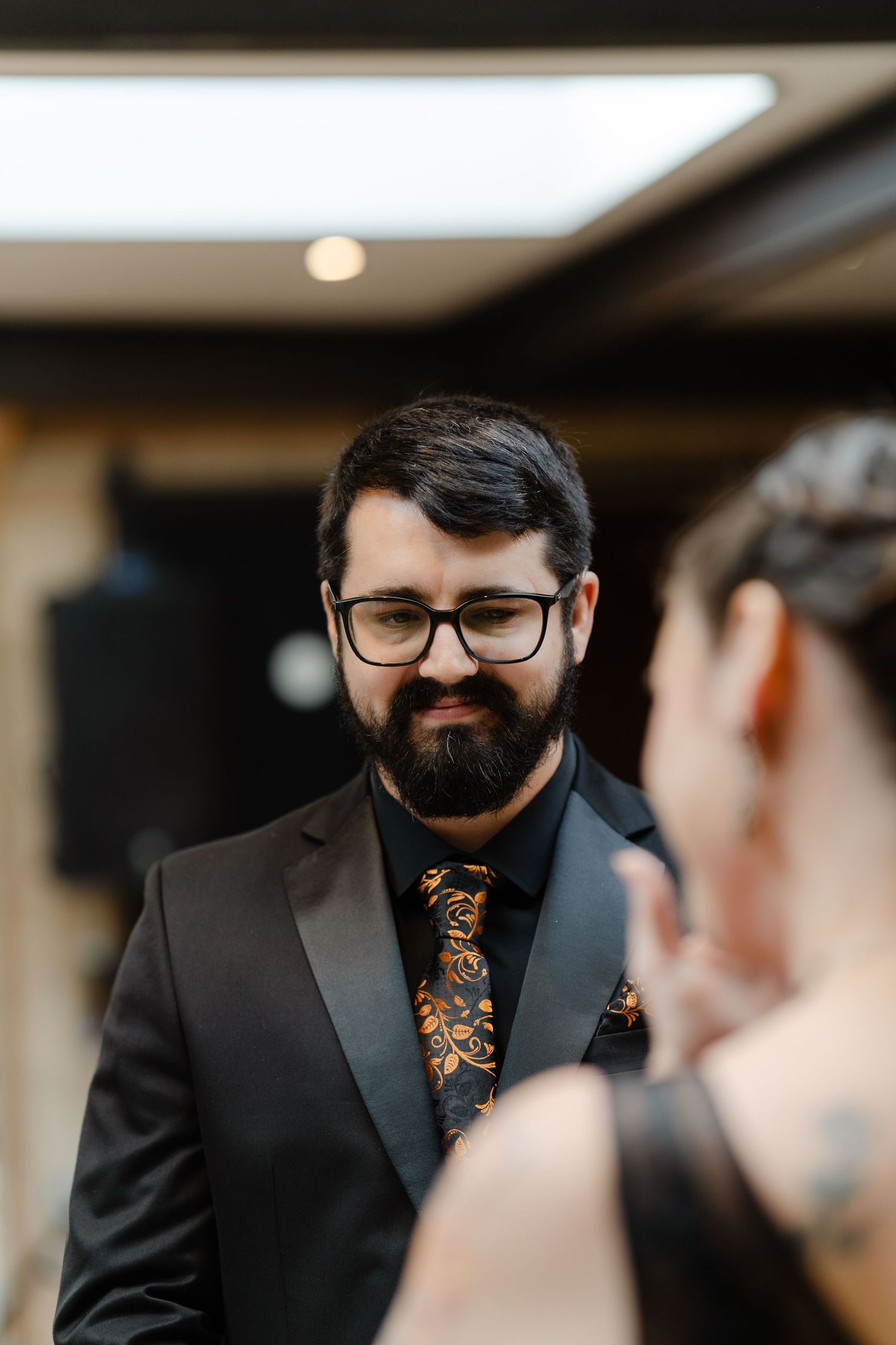 Bearded man in black suit with paisley tie and glasses smiling at someone during a formal event.
