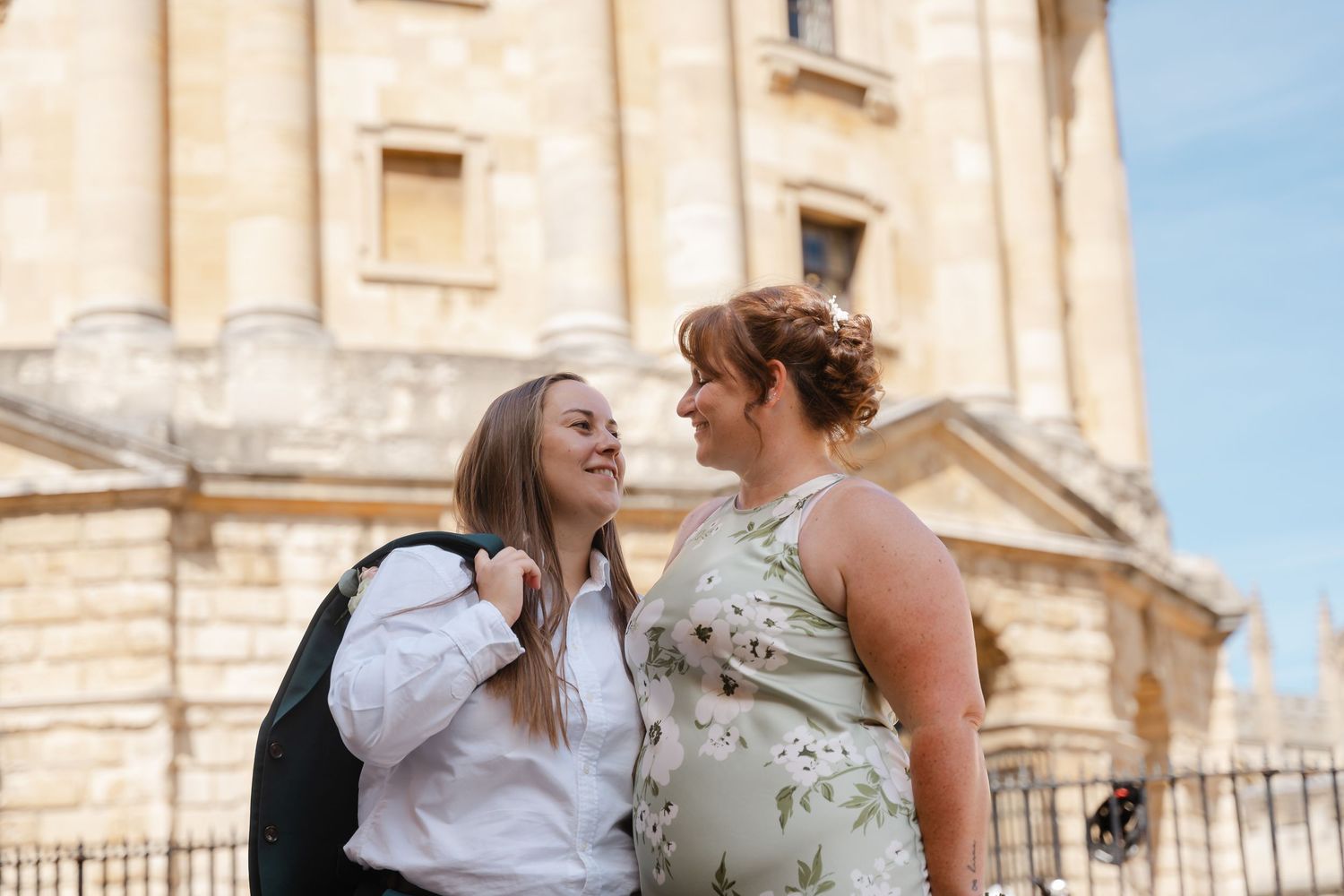 Two women smiling at each other near a historic stone building in Oxford on a sunny day.