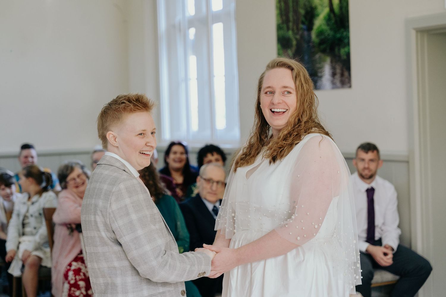 Two brides laughing joyfully during their wedding ceremony, one in a white dress and one in a plaid suit.