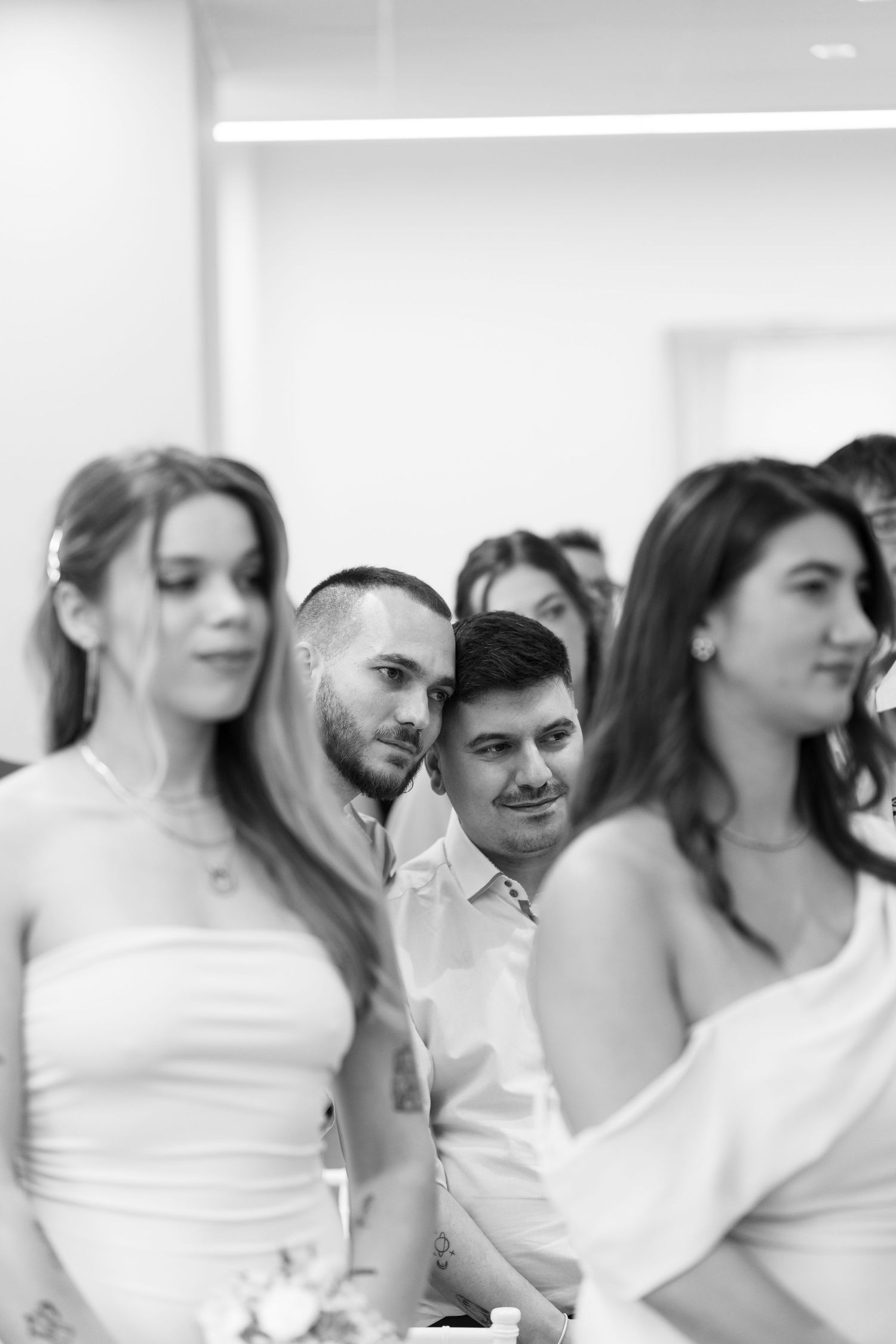 Black and white photo of wedding guests seated together, smiling and watching a ceremony in a bright indoor venue.