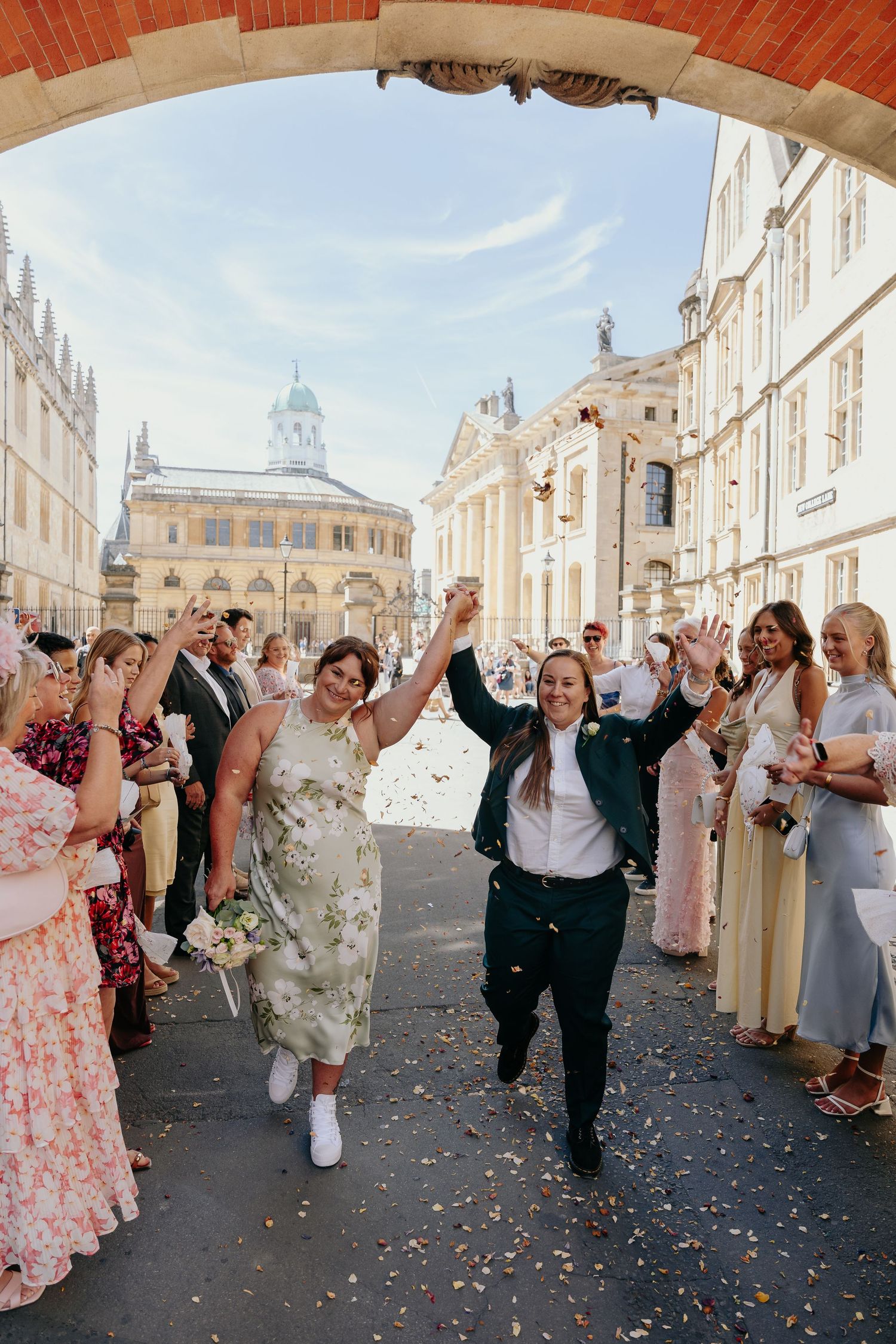 Two brides celebrate with confetti during their wedding exit under an arch in Oxford, England.