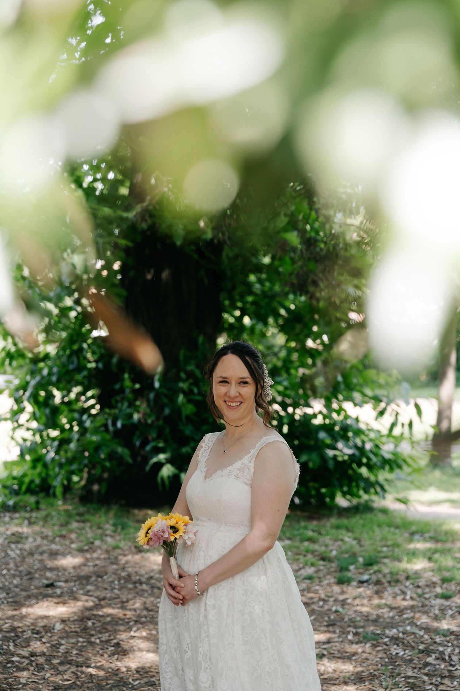 Smiling bride in white lace dress holding yellow bouquet, standing outdoors surrounded by lush green trees.