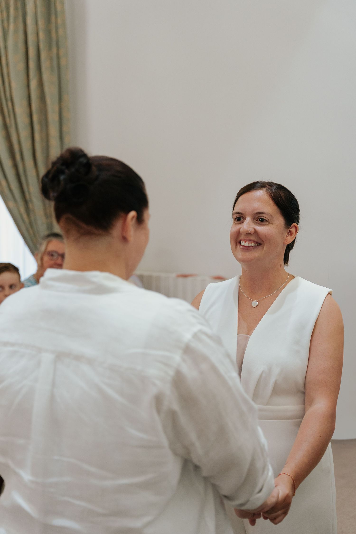 Two women in white attire hold hands and smile during an intimate wedding ceremony indoors.