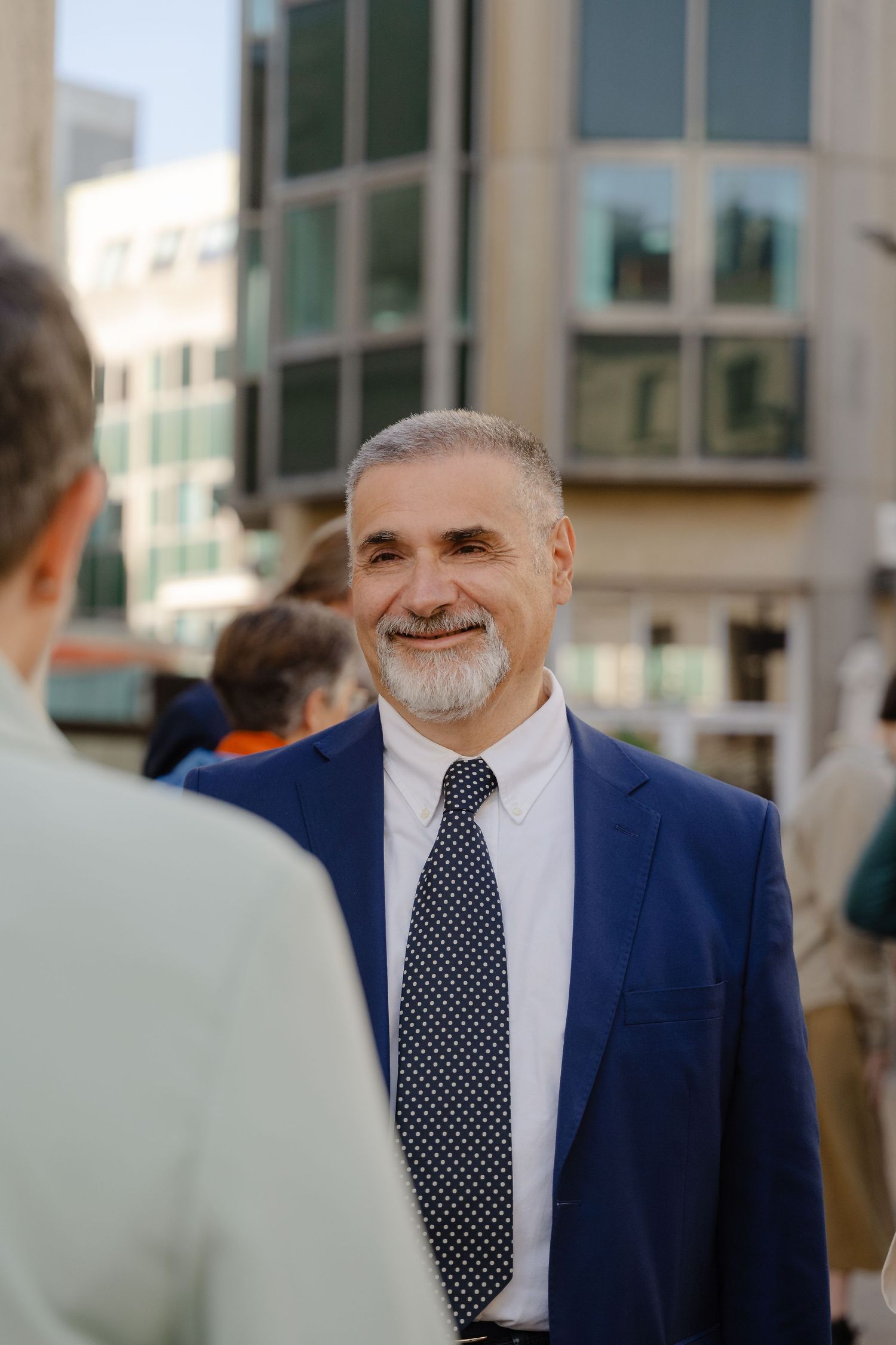 Smiling middle-aged man with gray beard wearing blue suit and polka dot tie outdoors near modern building.
