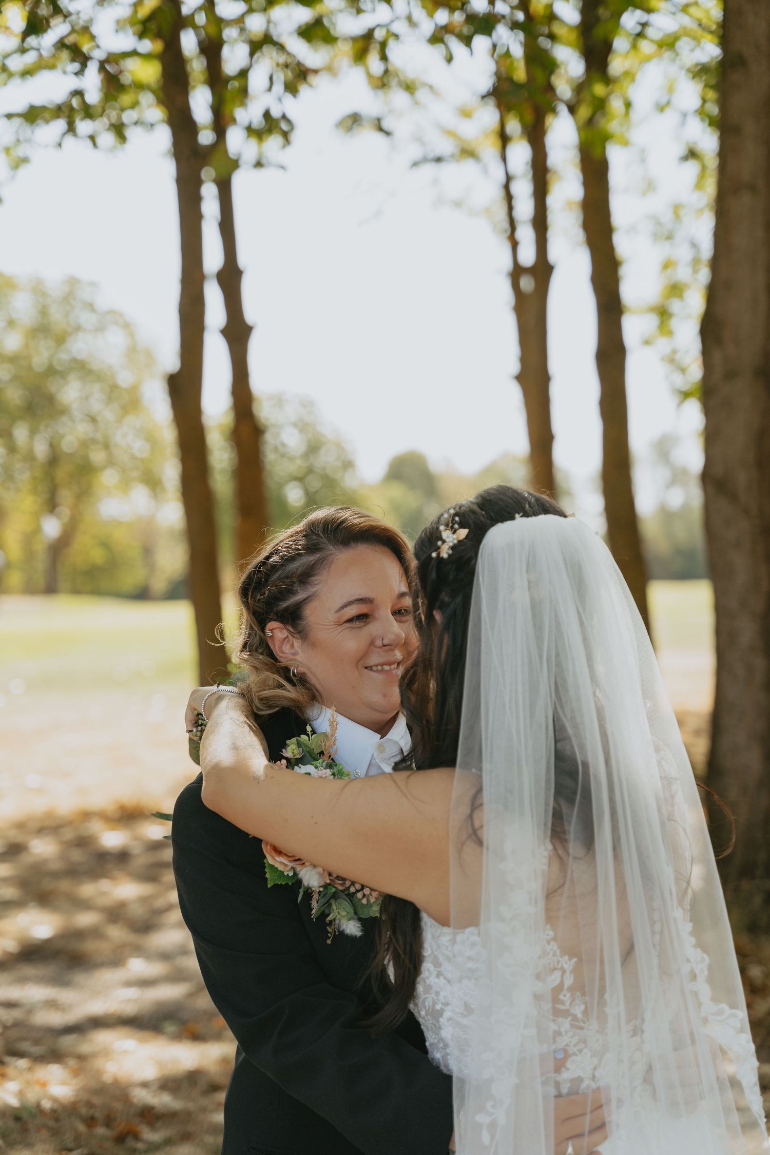 A bride in a white veil embraces a smiling woman in a black suit outdoors among trees on a sunny wedding day.