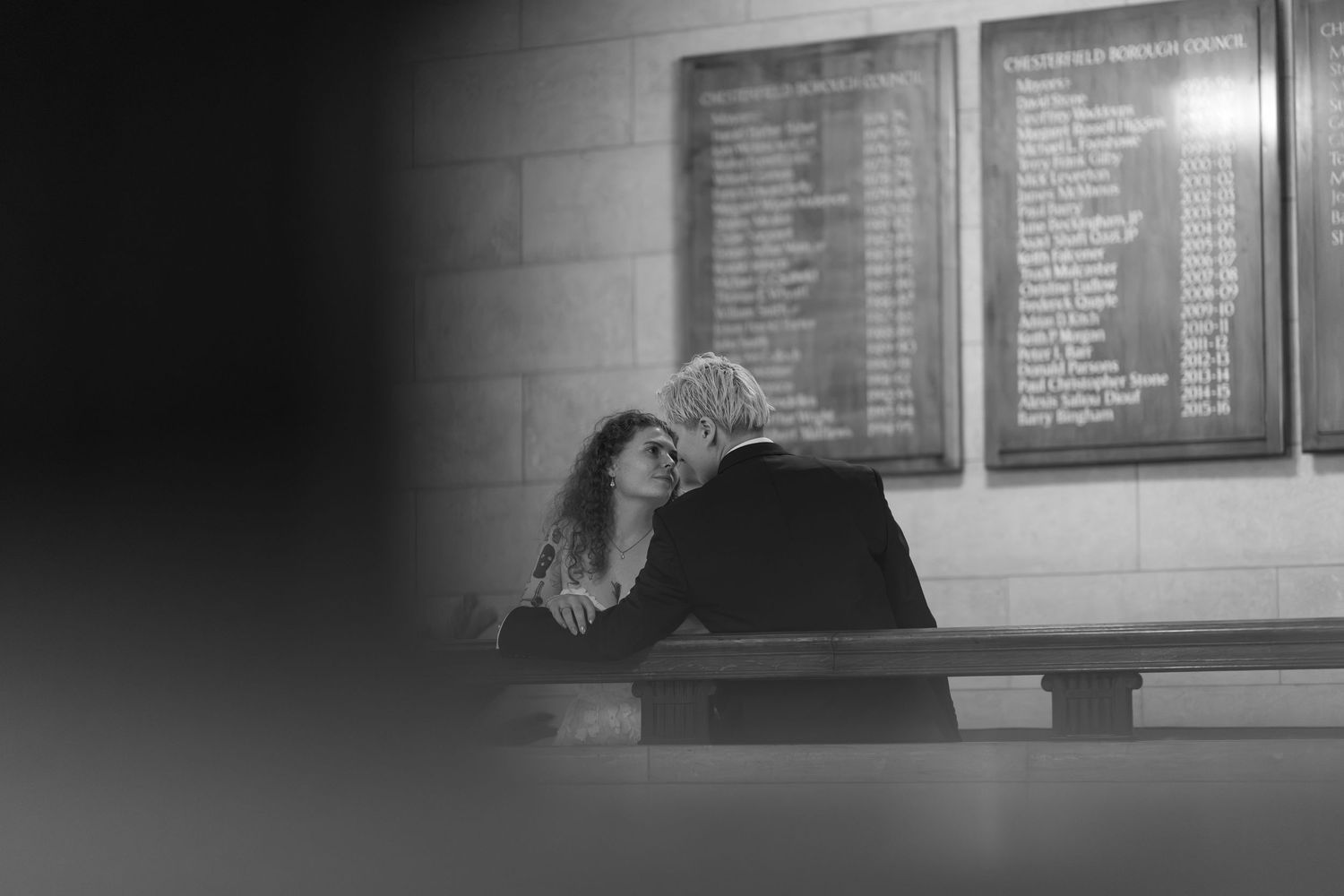Black and white photo of two people sitting close together on a bench beneath memorial boards.