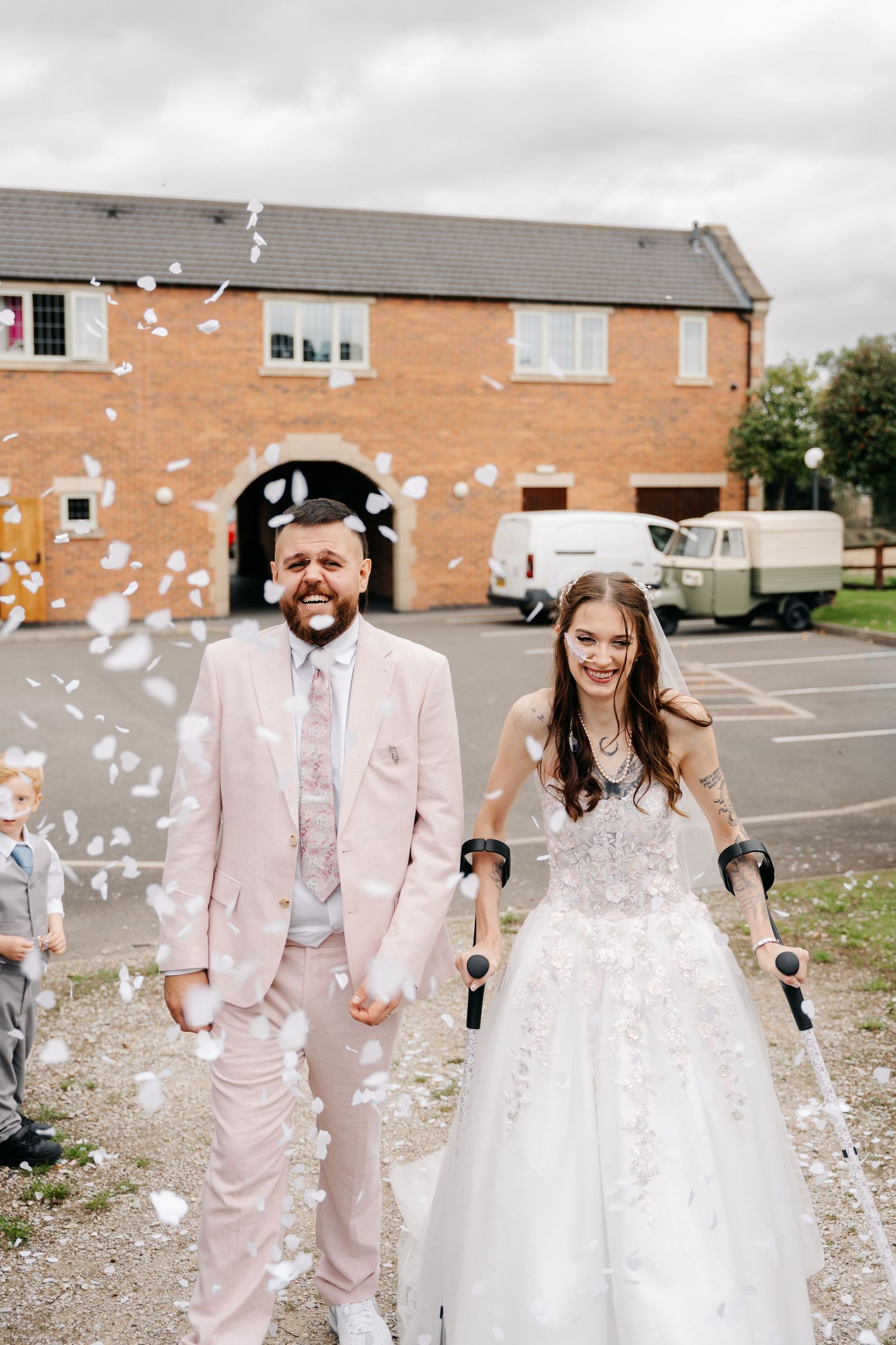 Bride with forearm crutches and groom in pink suit walk through white confetti outside a rustic brick wedding venue.