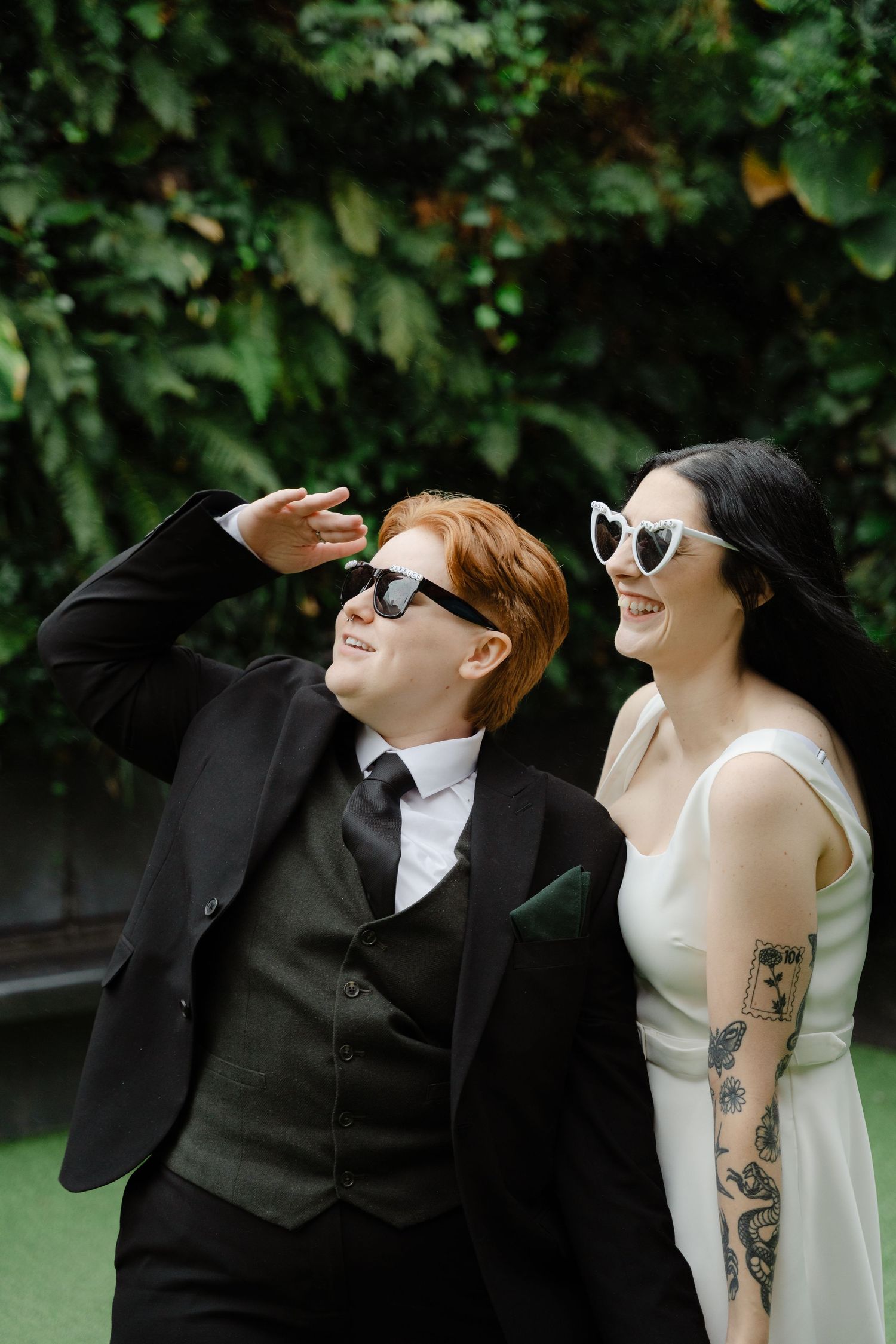 Two brides laughing outdoors, one in a black suit and one in a white dress, both wearing sunglasses on their wedding day.
