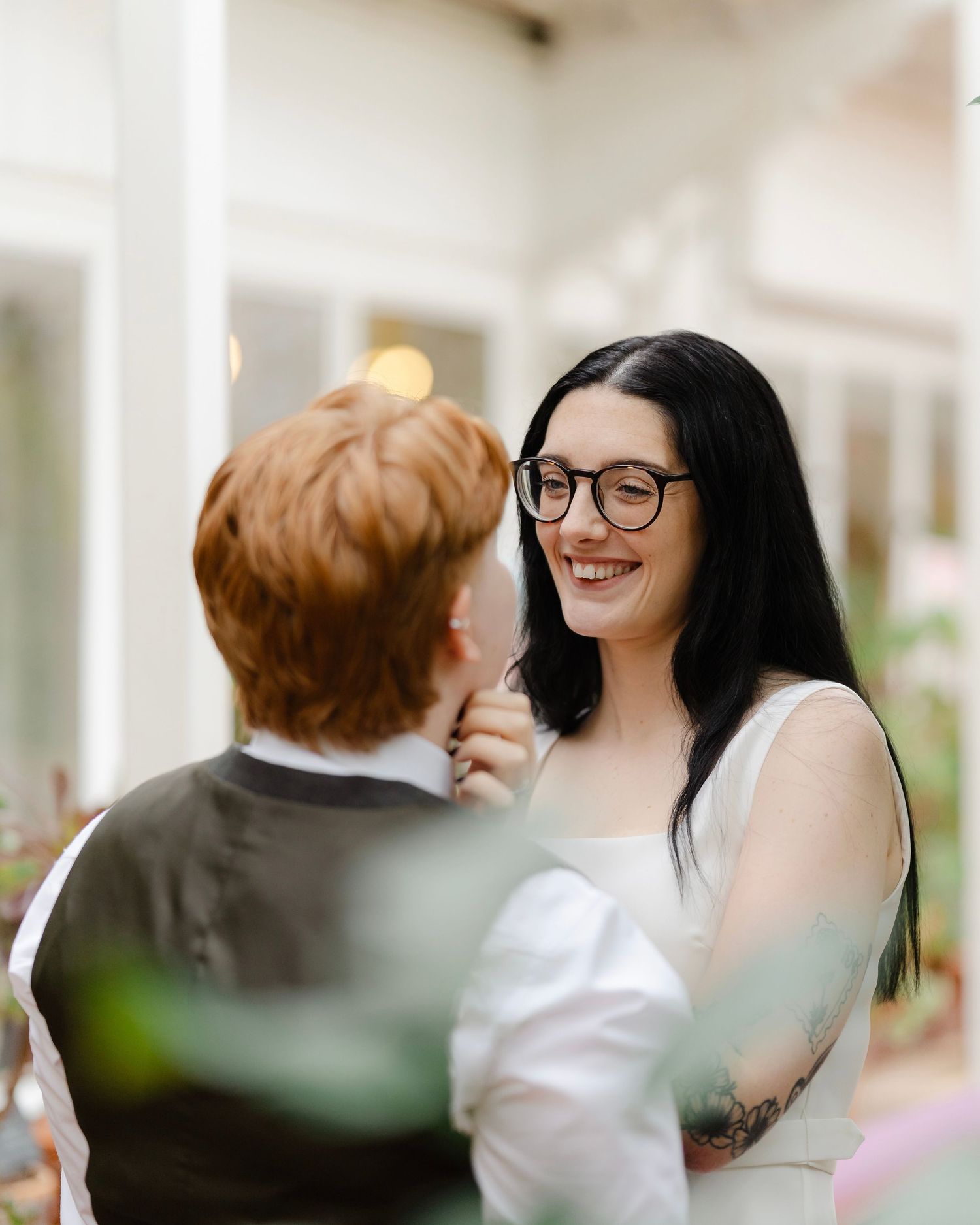 Two brides share a joyful moment, one with black hair and glasses, the other with red hair, at an outdoor wedding.