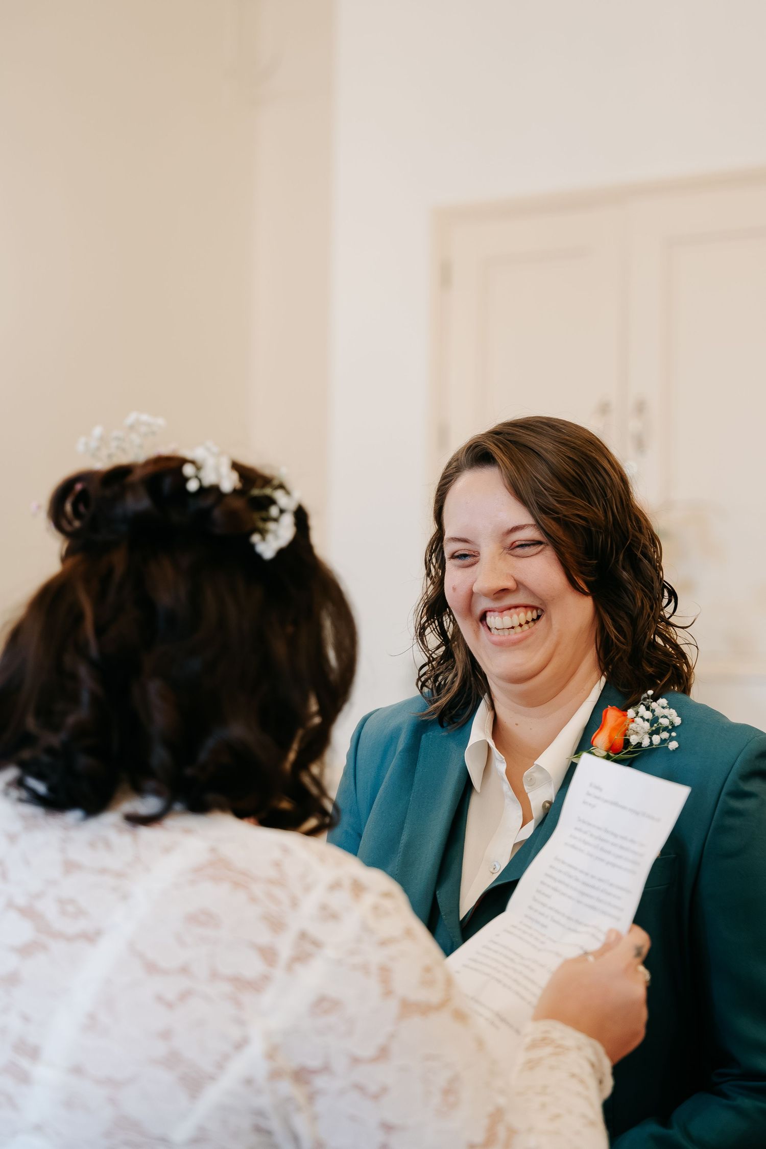 Two brides share a joyful moment during wedding vows, one in teal suit with boutonniere, other in lace dress with floral crown.