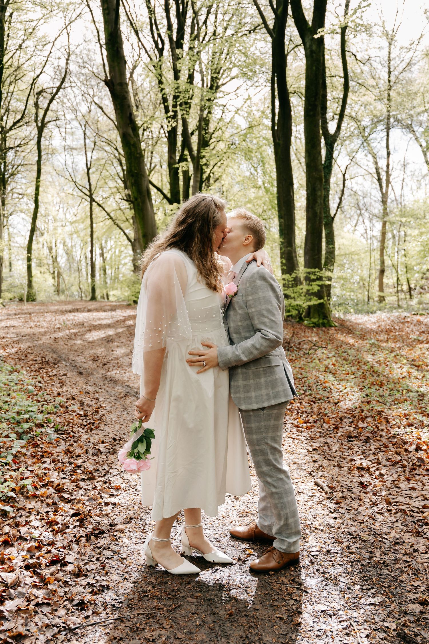 A couple shares a romantic kiss on a forest path, bride in white dress holding bouquet, groom in plaid suit.