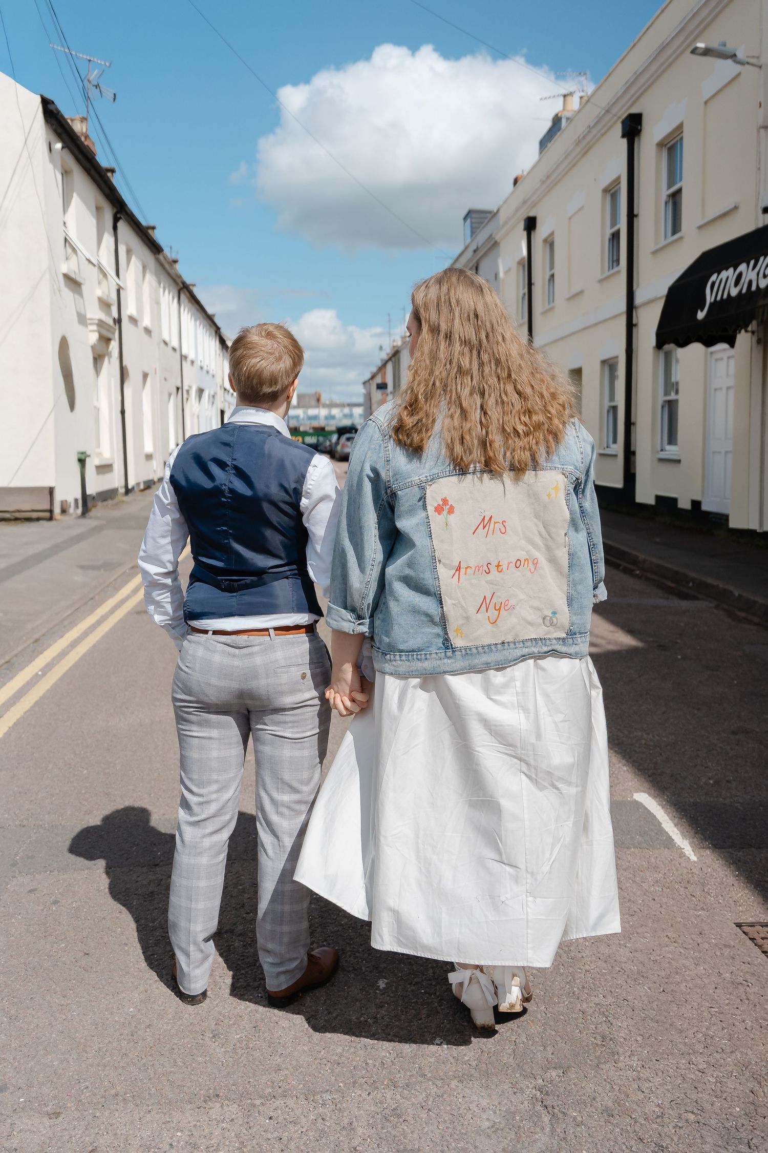 Newlywed couple walking hand in hand down a sunny street, bride wearing a denim jacket with Mrs written on the back.