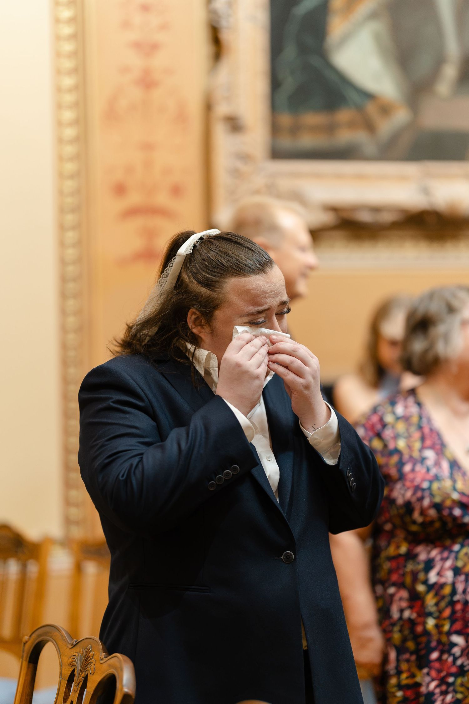 Emotional guest in navy suit wiping tears with handkerchief during wedding ceremony in ornate venue.