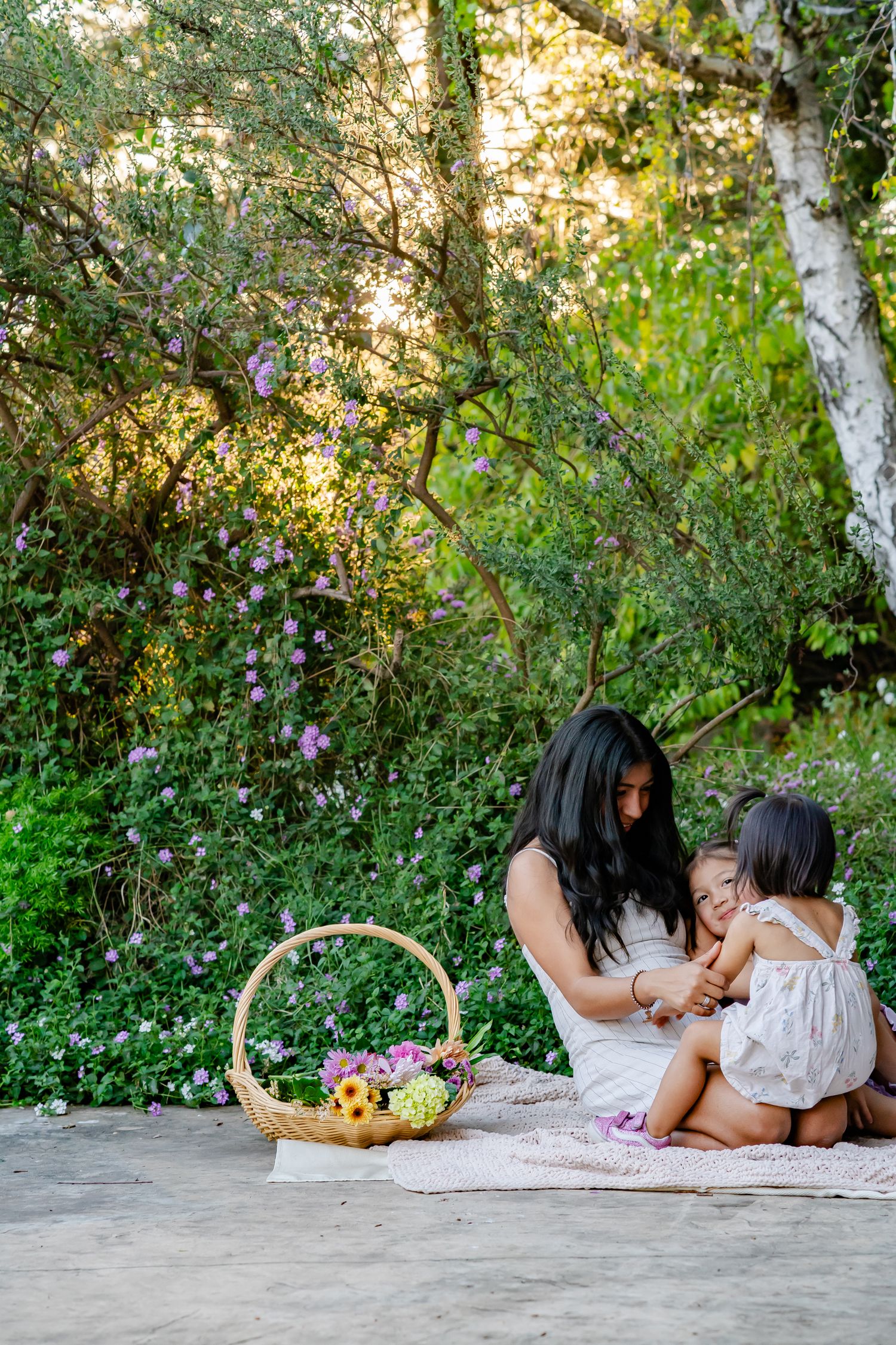mother snuggles her daughters surrounded by forest, flowers, greens, and a basket of flowers