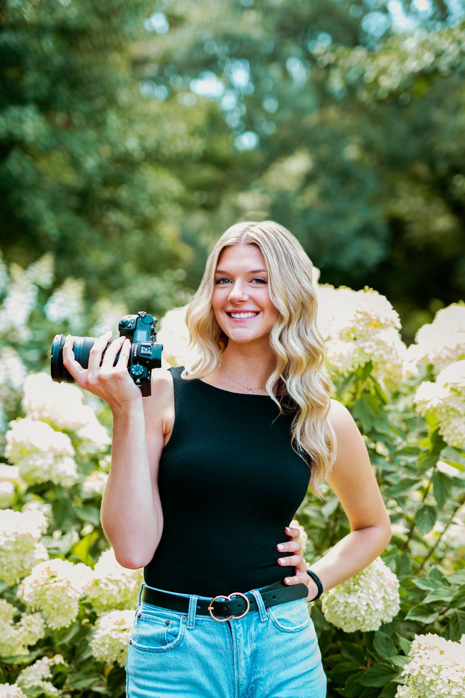 Photographer with wavy blonde hair holding camera in black tank top and jeans poses by blooming hydrangea bushes.