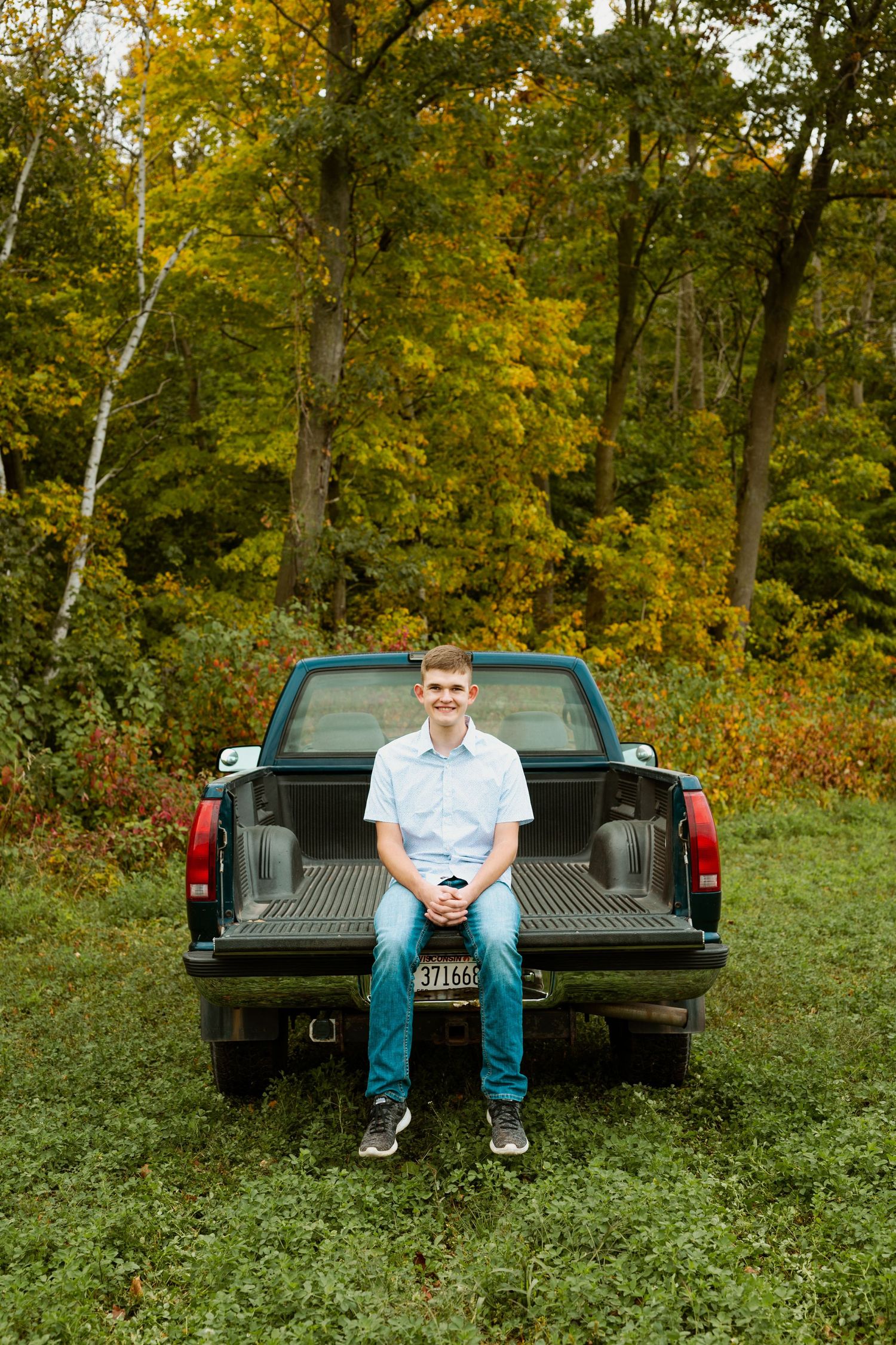 Student sits on tailgate of pickup truck surrounded by autumn foliage in outdoor senior portrait session.